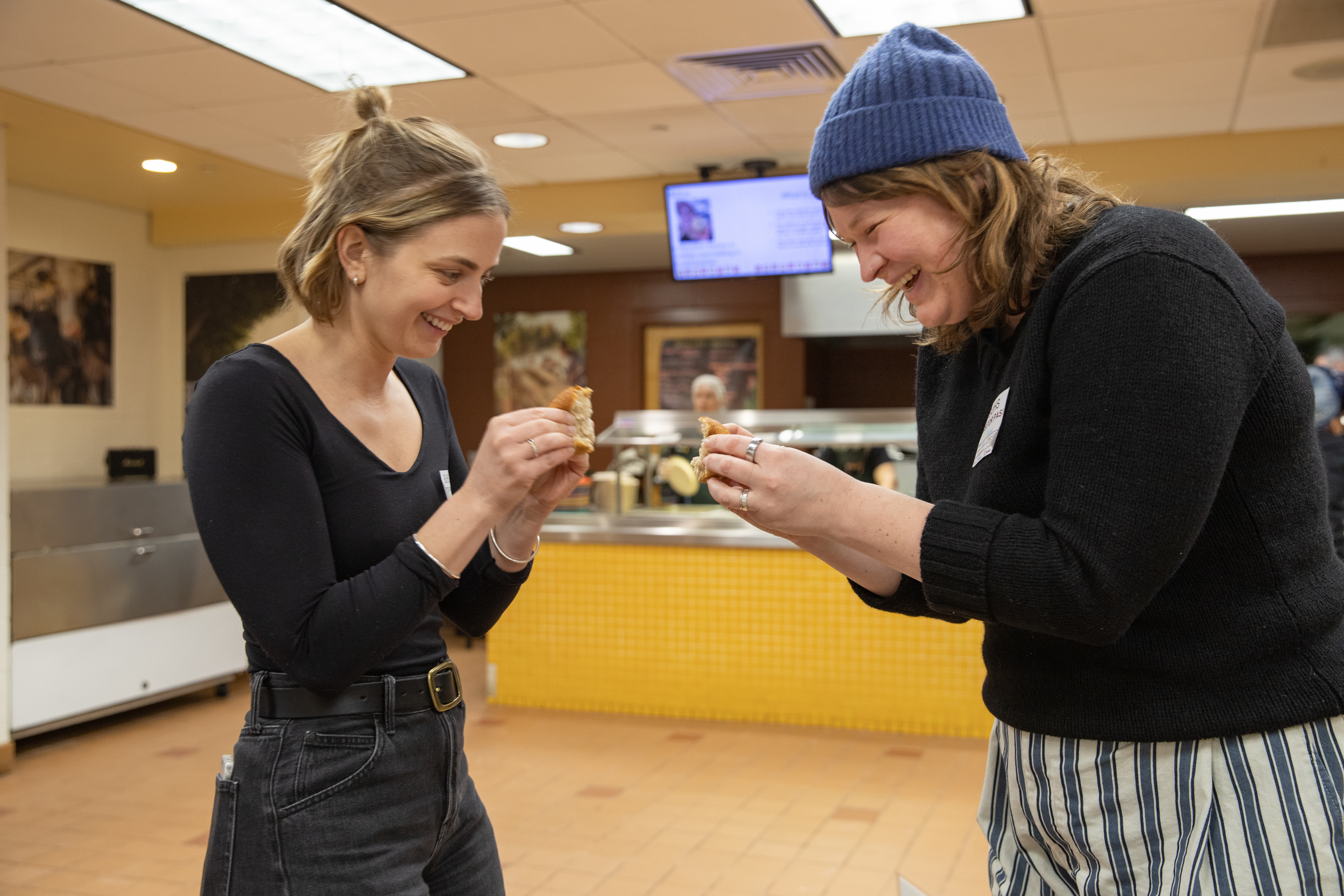 NOFA-VT Farm to Institution Program Director Kayla Strom and NOFA-VT Farm to School Local Procurement Specialist Becca Perrin are seen sharing an smile and tapping their local bread rolls together for a high-five in the Brattleboro school cafeteria