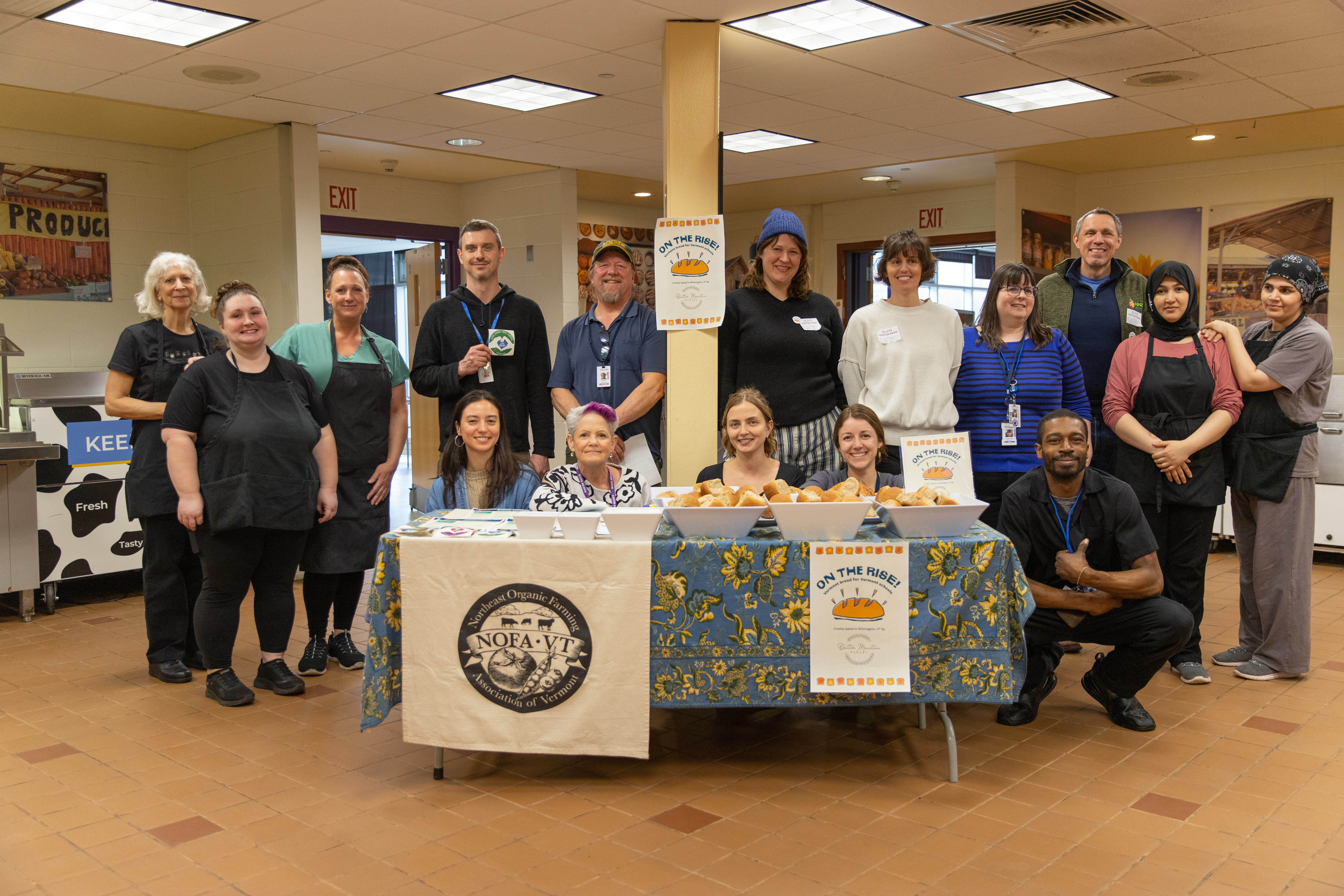 A group photo of the school nutrition professionals, NOFA-VT's farm to school and farm to institution team, and the team from VTFEED stand smiling in the Brattleboro school cafeteria