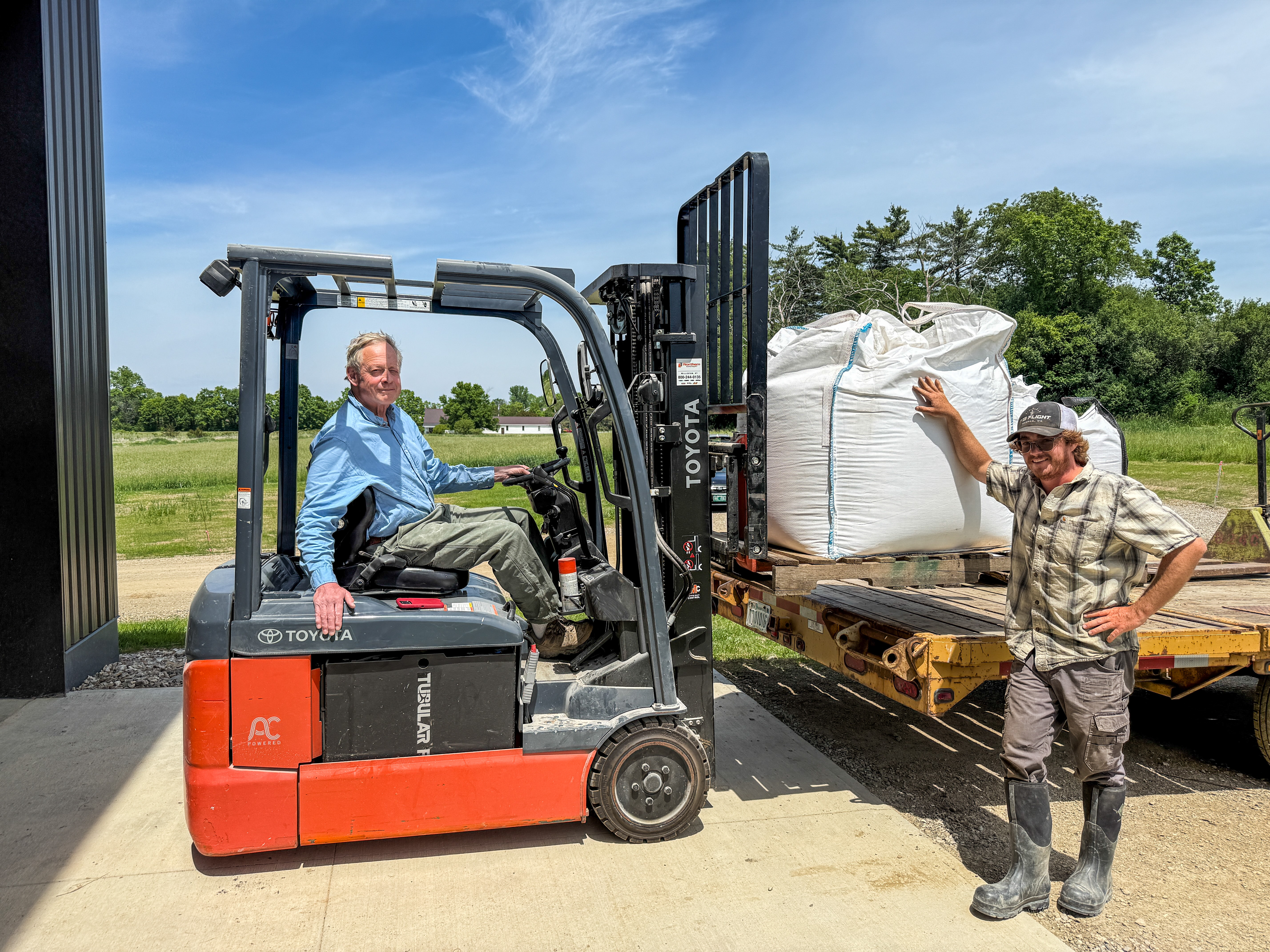 Todd Hardie of Champlain Valley Grain Center is shown operating a forklift that is receiving a bulk order of organic grain grown by Tyler of Granite Grains, who stands proudly beside the large sack of grain on the forklift.
