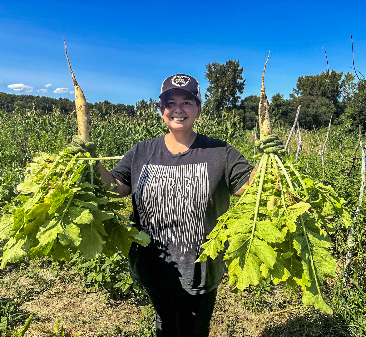 NOFA-VT Board Member Alexis Yamashita is pictured in a farm field holding a large daikon radish upside down in each hand as she stands smiling betwen them.