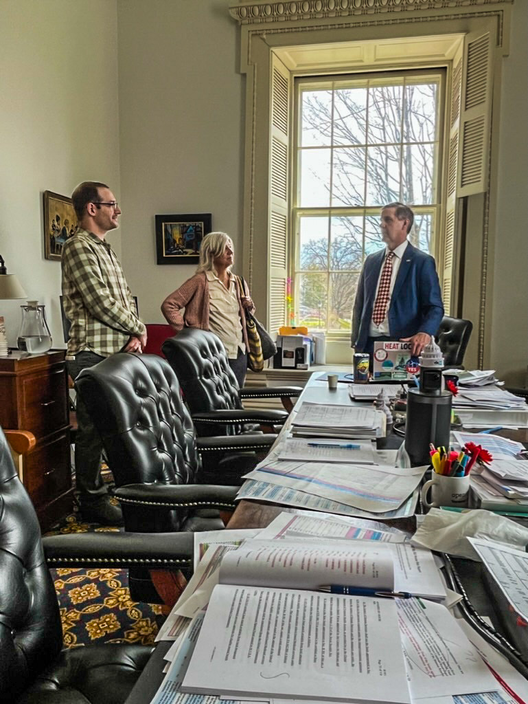 NOFA-VT Board Member Nick Sibley and NOFA-VT Finance Director Donna Samson are pictured chatting with Andy Perchlik, Chair of Senate Appropriations, in an office in the State House at the end of a long table covered with papers. 