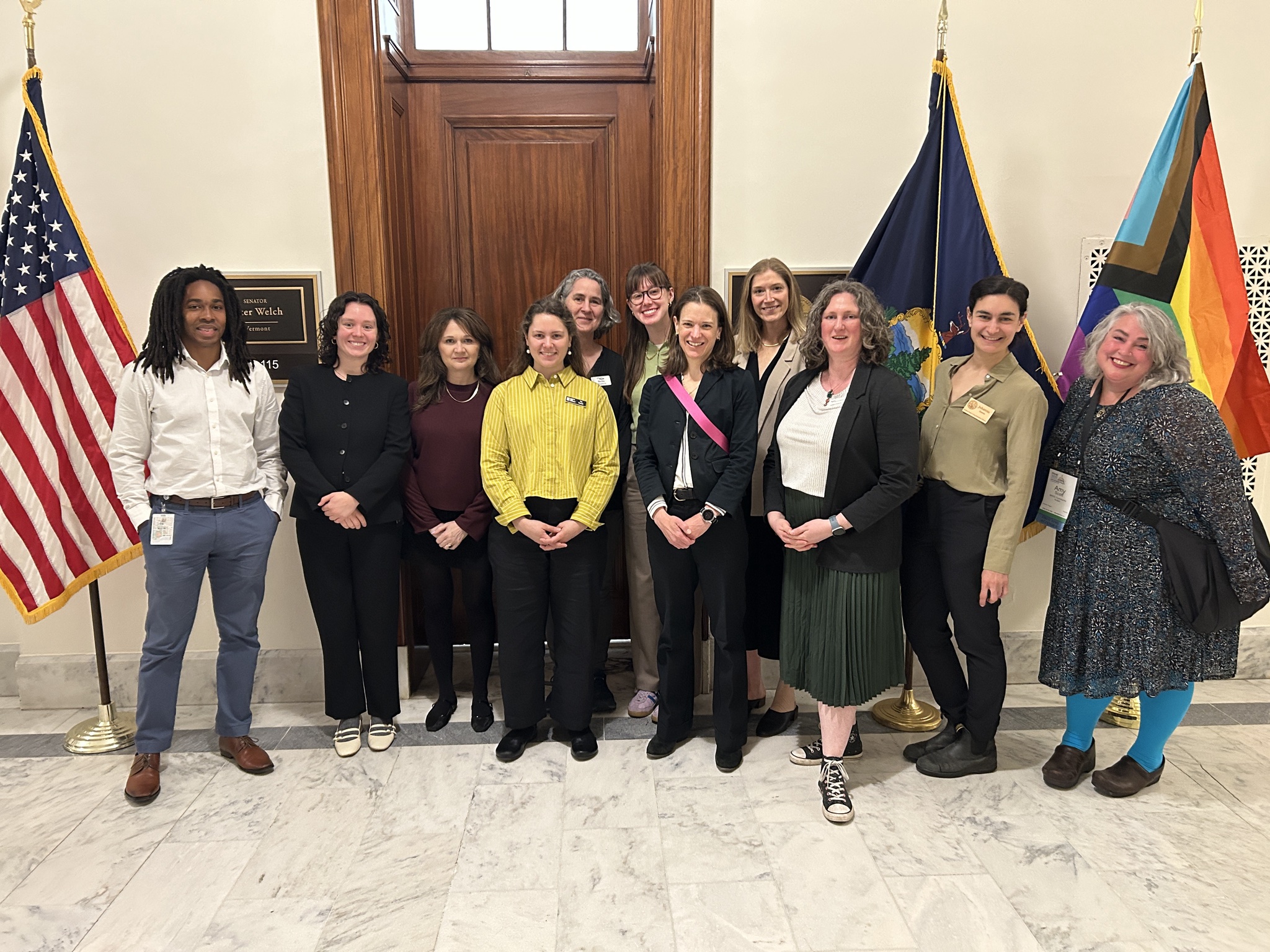 Members of the Vermont Food Security Coalition gathered in DC for the National Anti-Hunger Policy Conference. They are standing outside a Representative Becca Balint's office and are flanked by a US flag, a VT flag, and a pride flag