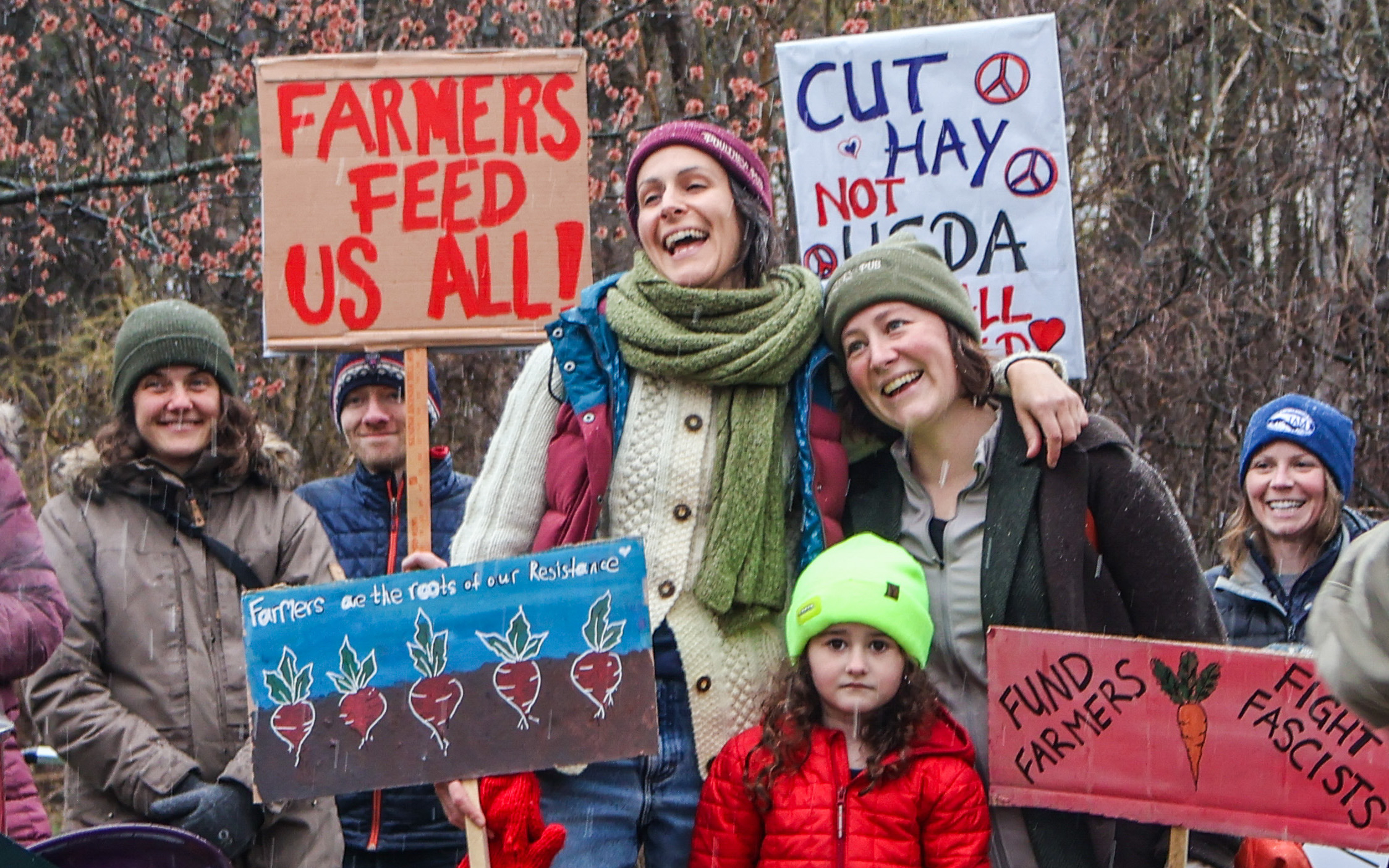 Farmers, farmworkers, and food system advocates stand in a crowd with signs reading "Farmers feed us all!," "Cut Hay, not USDA," "Farmers are the roots of our resistance," and "Fund Farmers, Fight Fascists"