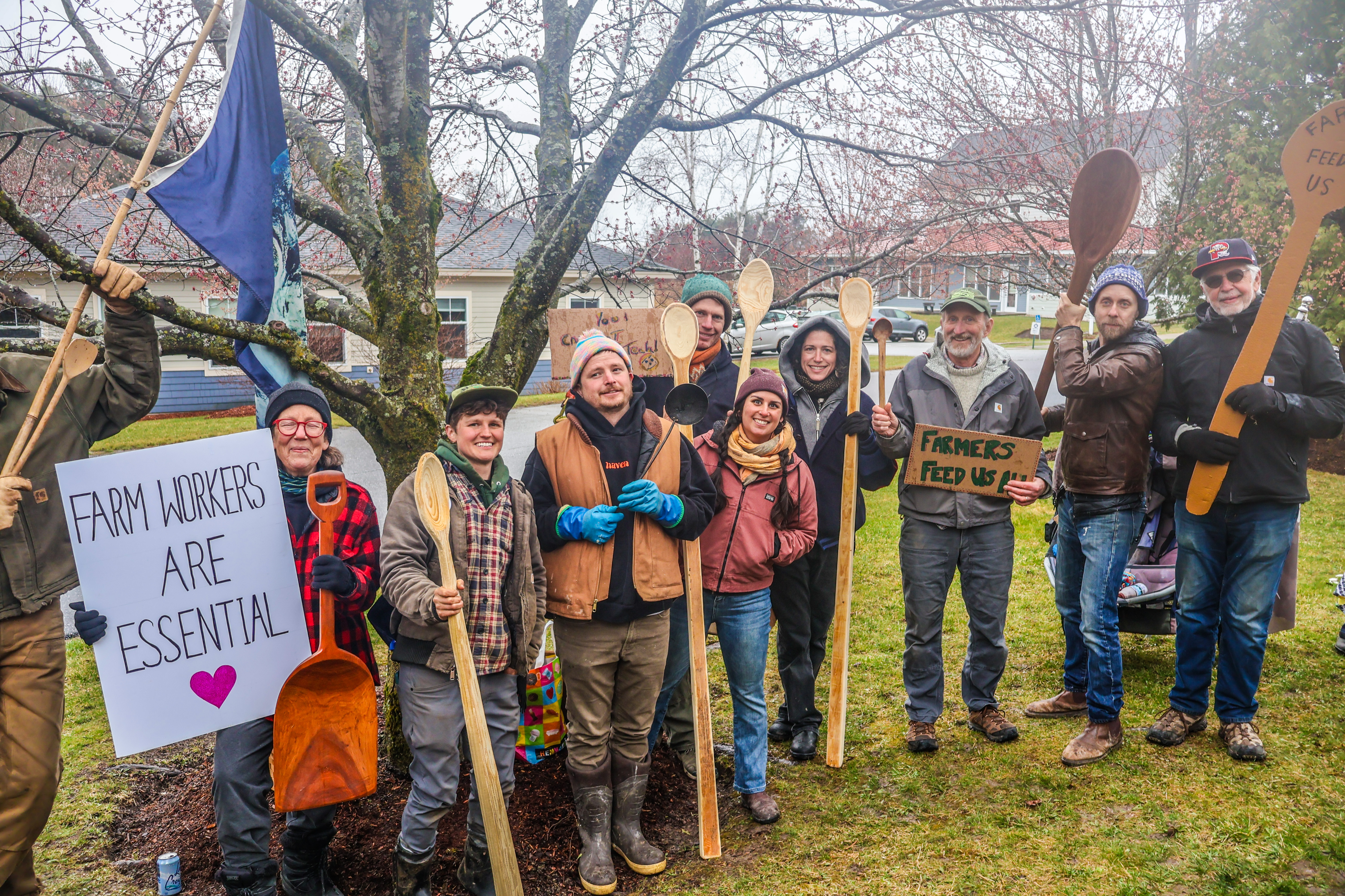 A group of NOFA-VT members, farmers, and farmworkers holding giant spoons and signs reading "Farmworkers Are Essential" and "Farmers Feed Us All" at a NOFA-VT action at the Middlebury USDA offices