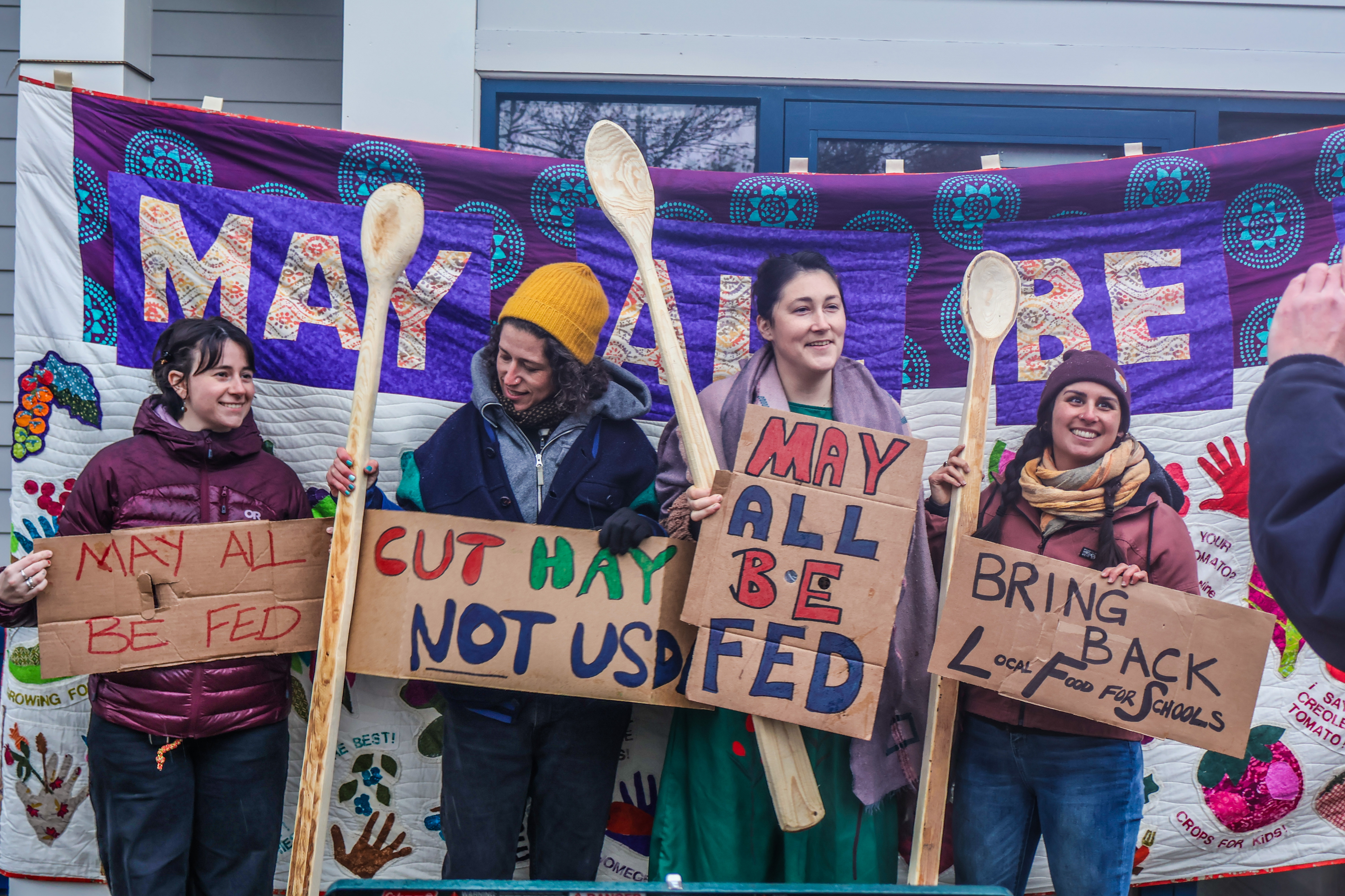 Farmers and food system advocates stand holding long-handled spoons and signs reading "May all be fed," "cut hay, not USDA," and "Bring back local food for schools" standing in front of a large banner reading "May All Be Fed"