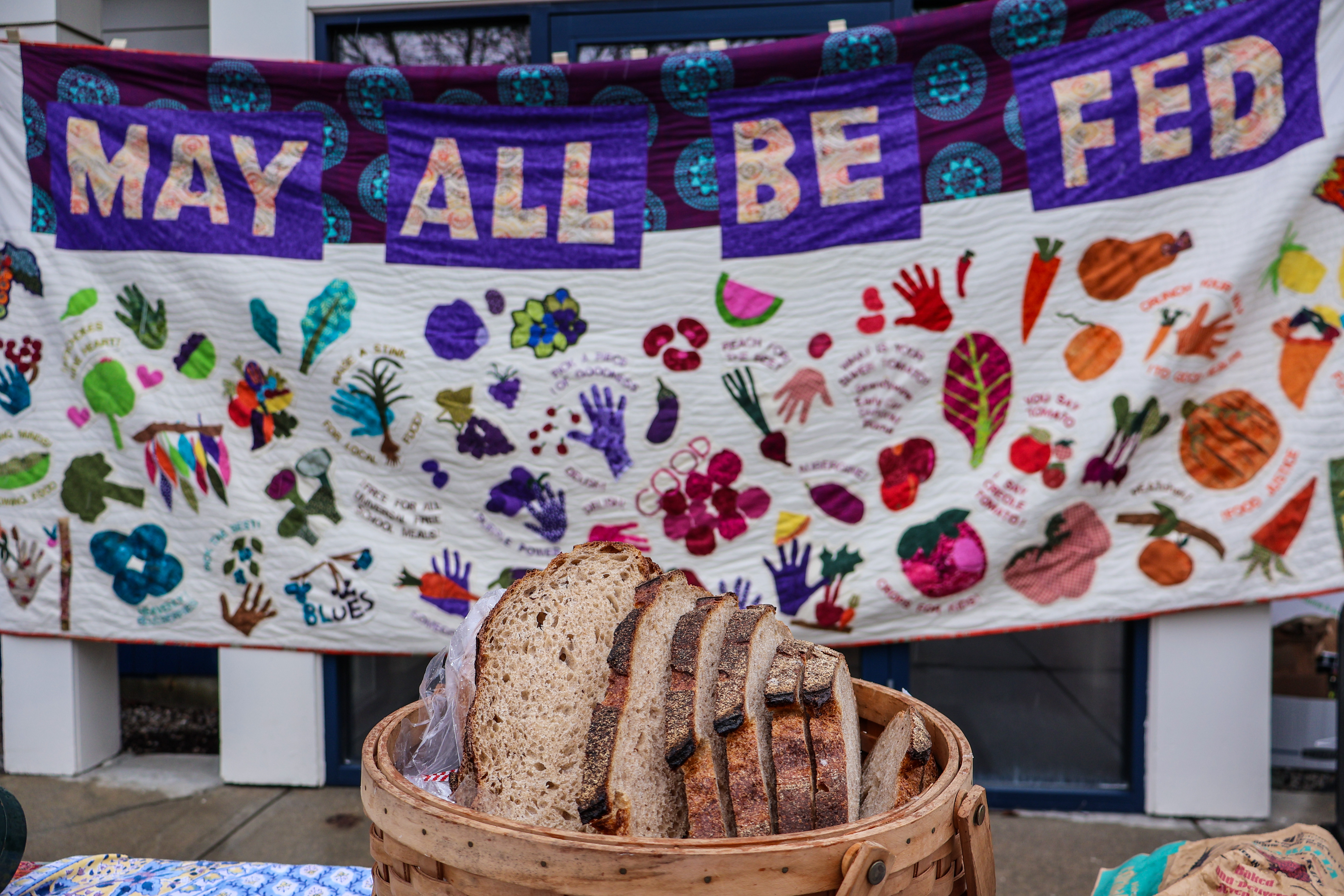 A fresh loaf of sliced artisan bread on a table with a banner behind it reading "May All Be Fed"
