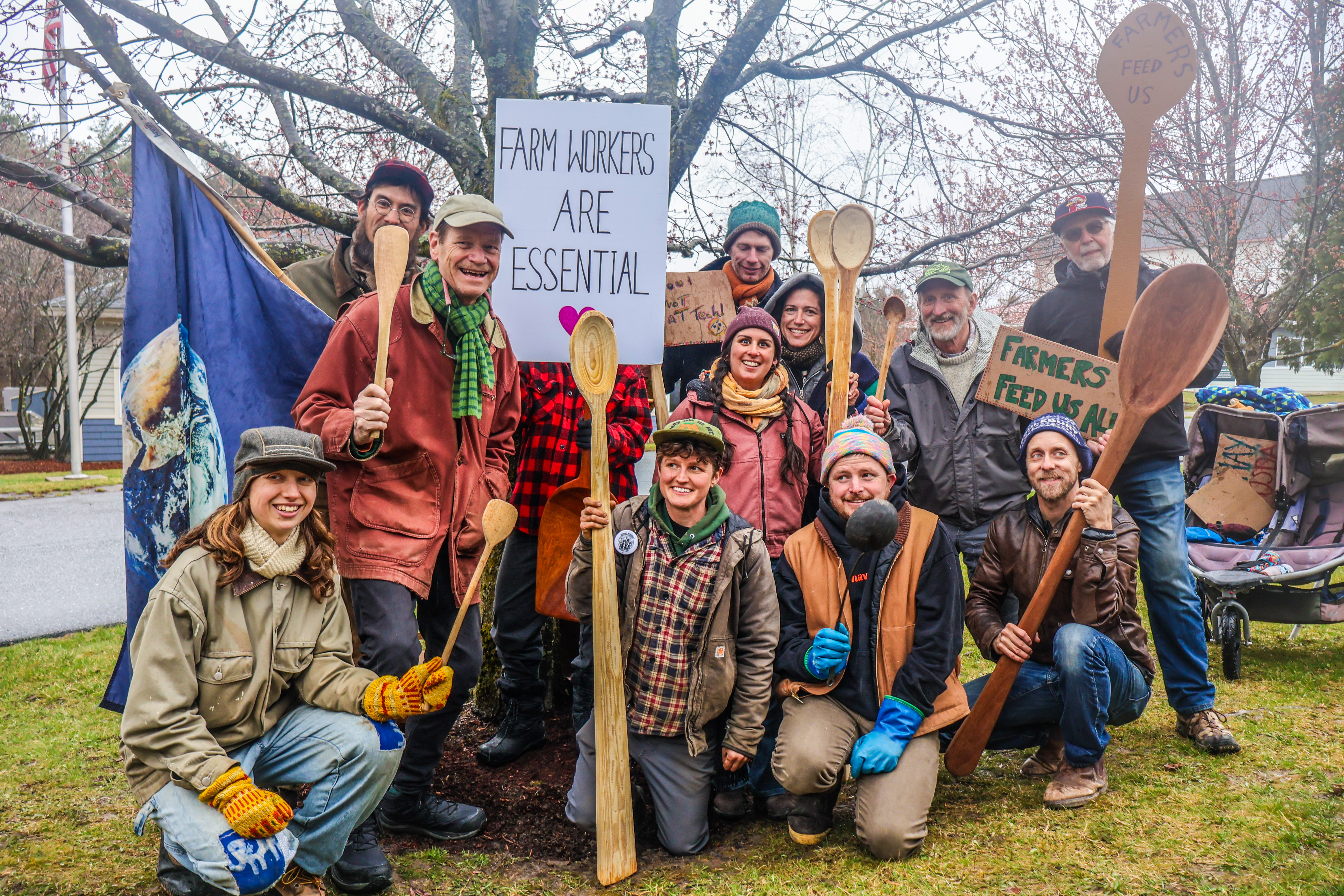 A large smiling group of farmers, farmworkers, and food system advocates stand holding long-handled spoons and signs reading "farmworkers are esential" and "farmers feed us all" at a NOFA-VT Farmer Day of Action at the USDA offices