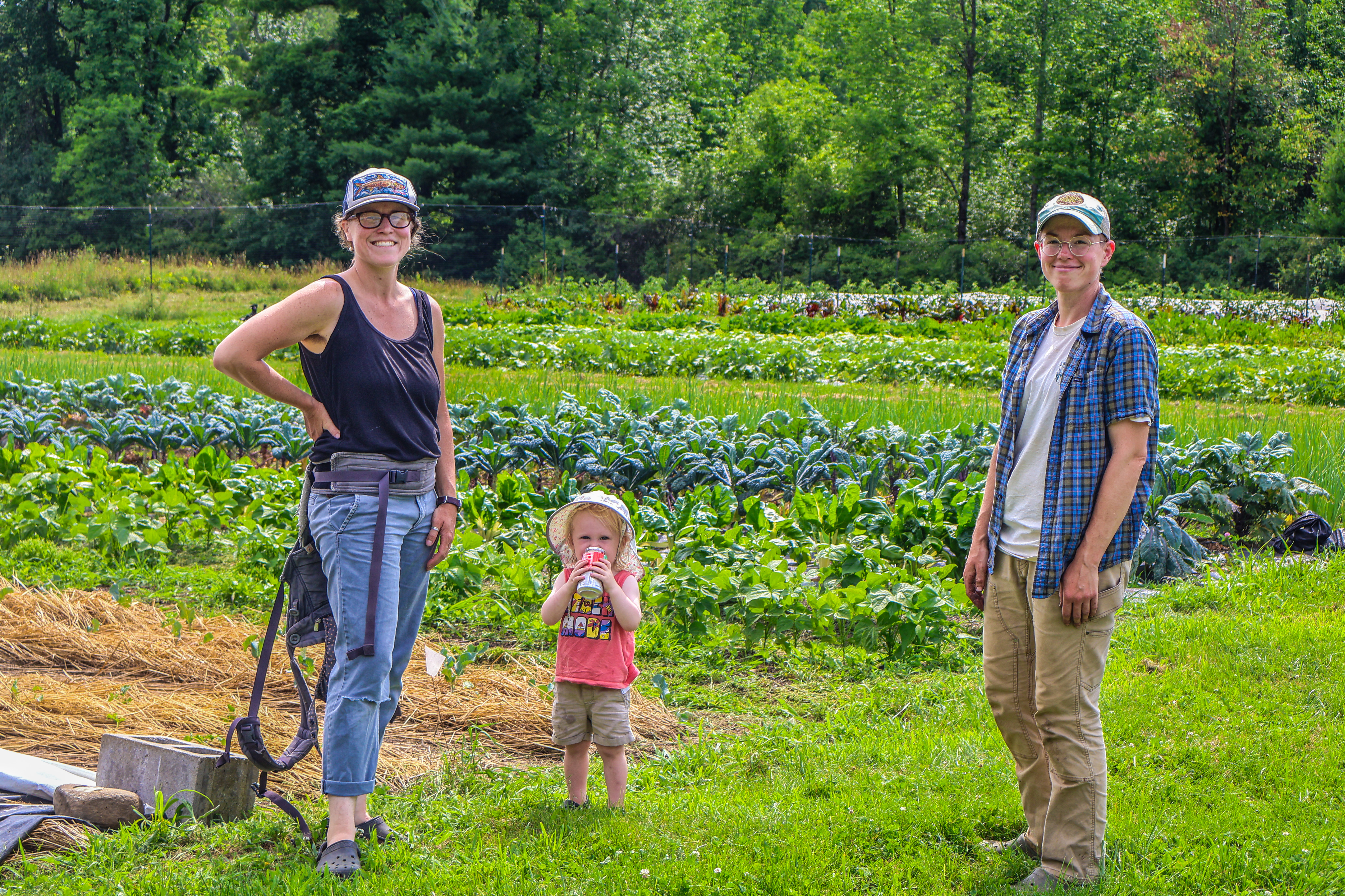 Resilience Grantee Paige Wener stands with her hand on her hip with her farm fields in the background and her kiddo and farm staff member stand to her left. 