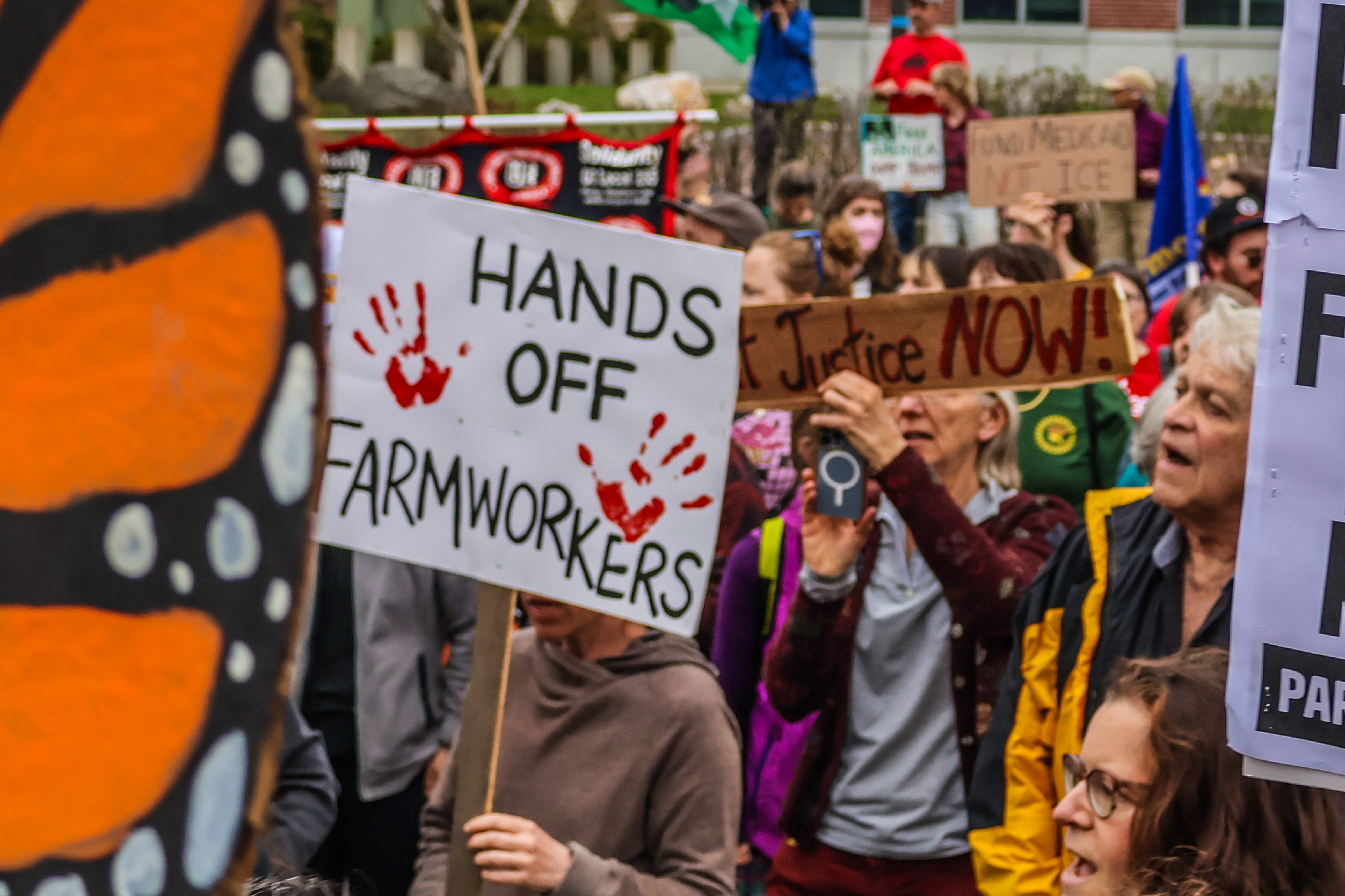 large crowd of protesters at a May Day rally in Burlington, centering a sign reading "Hands Off Farmworkers"