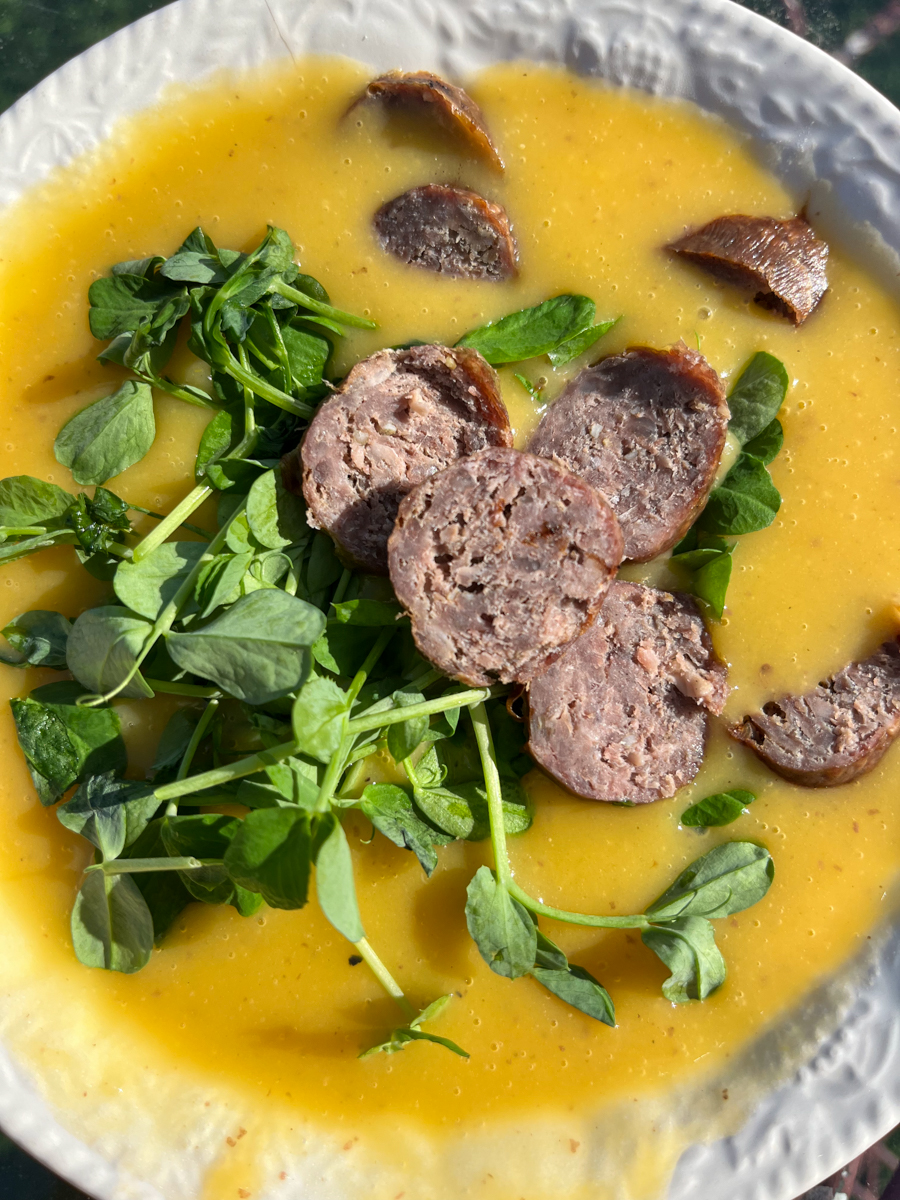 A bowl of yellow soup with microgreens and sausage as toppings