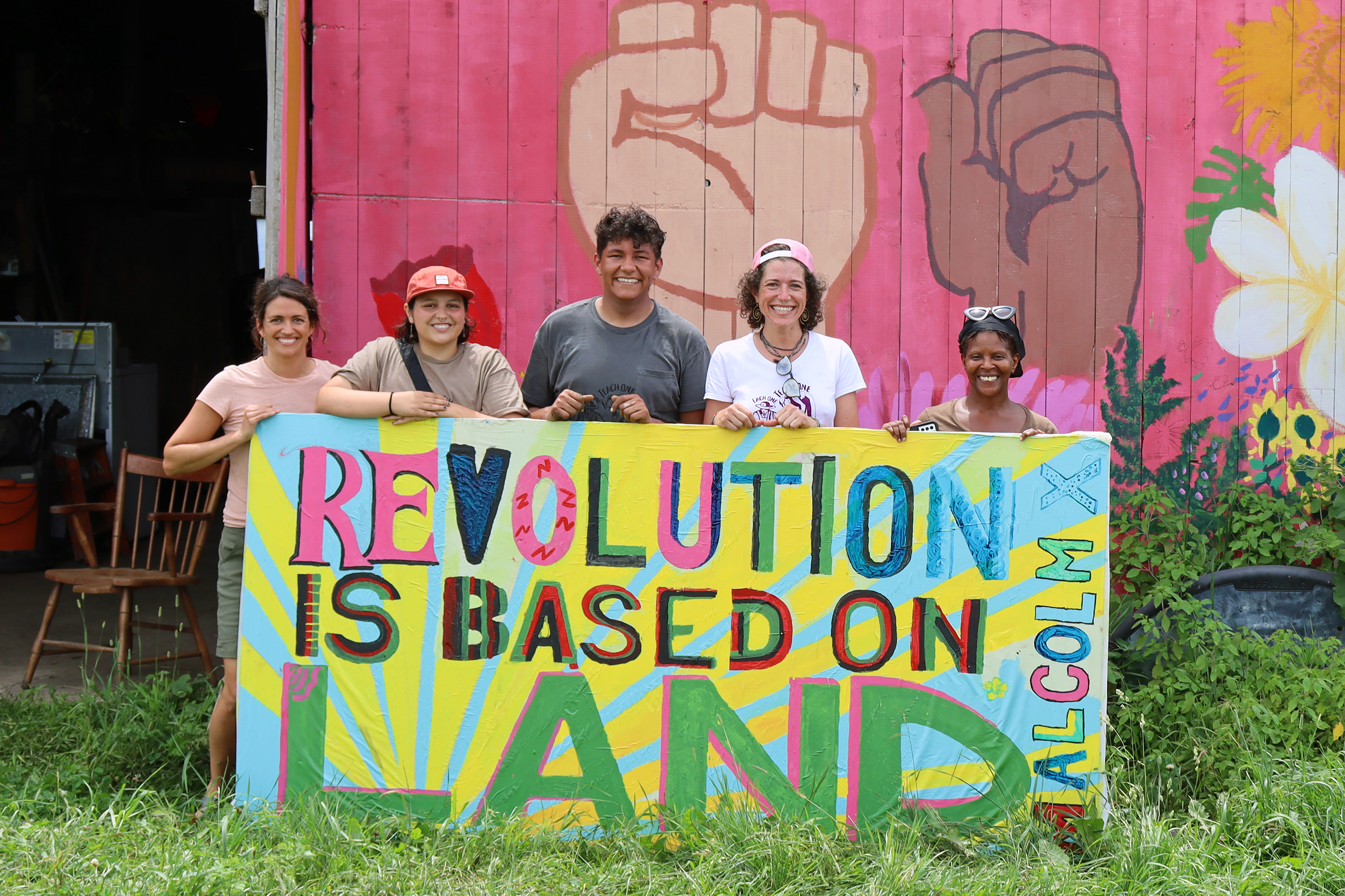 A group of people at an Agroecology workshop honoring peasant farming wisdom standing behind a sign reading "Revolution is Based on Land" and with a mural on the barn behind them of fists in the air and colorful flowers