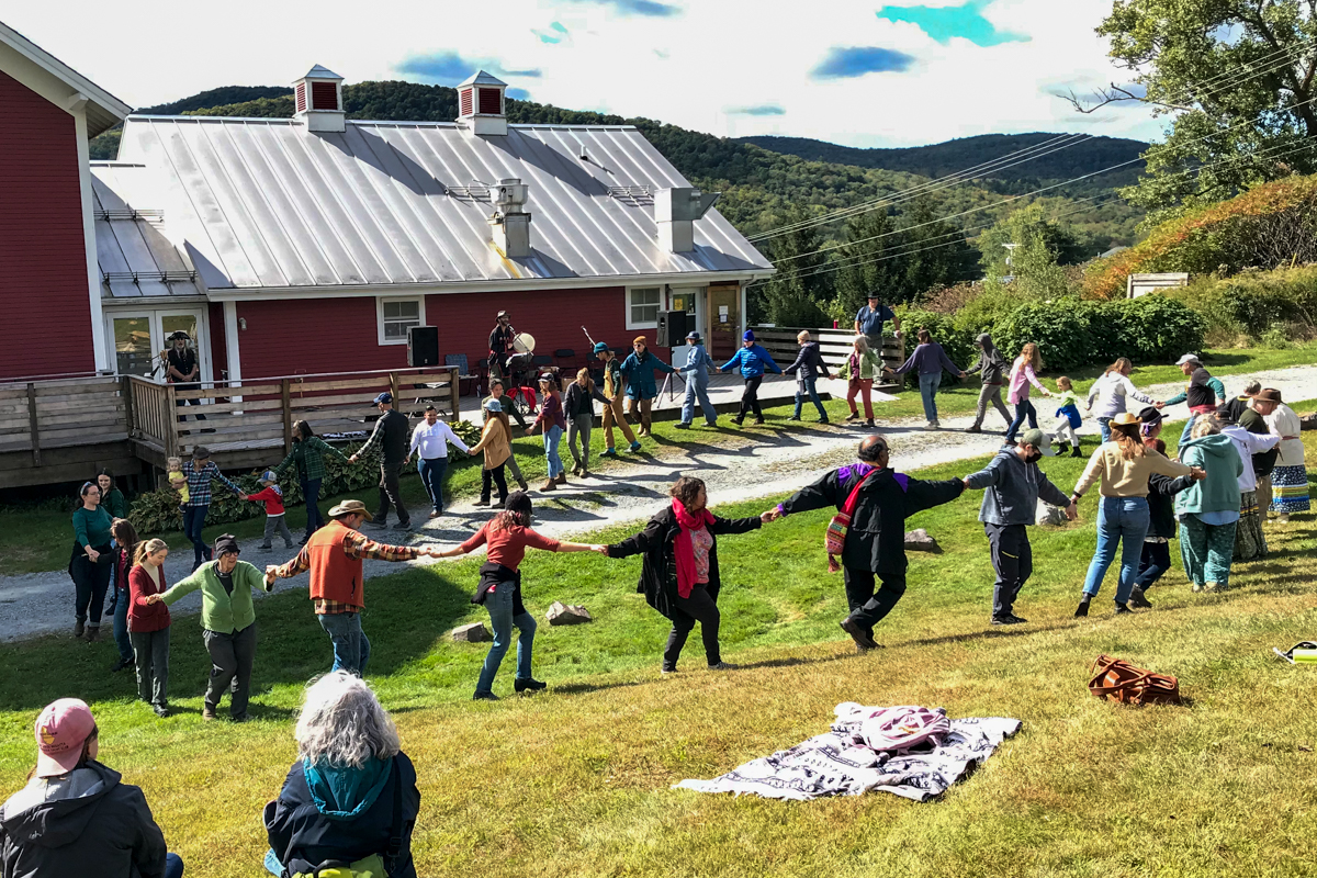 A group of roughly 20 people hold hands in a big circle in front of a barn