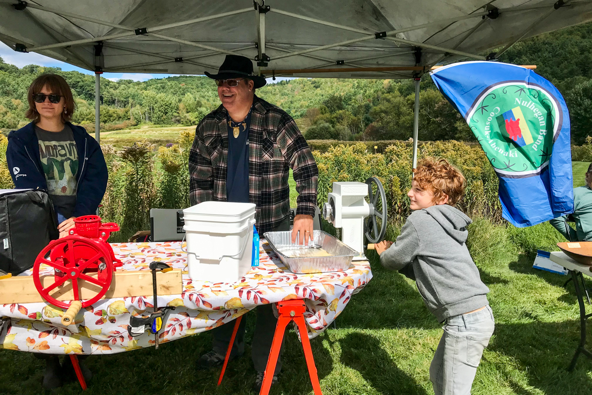 Two people stand behind a table at a tent while a kid cranks a mid-sized metal tool that is processing cobs of corn.