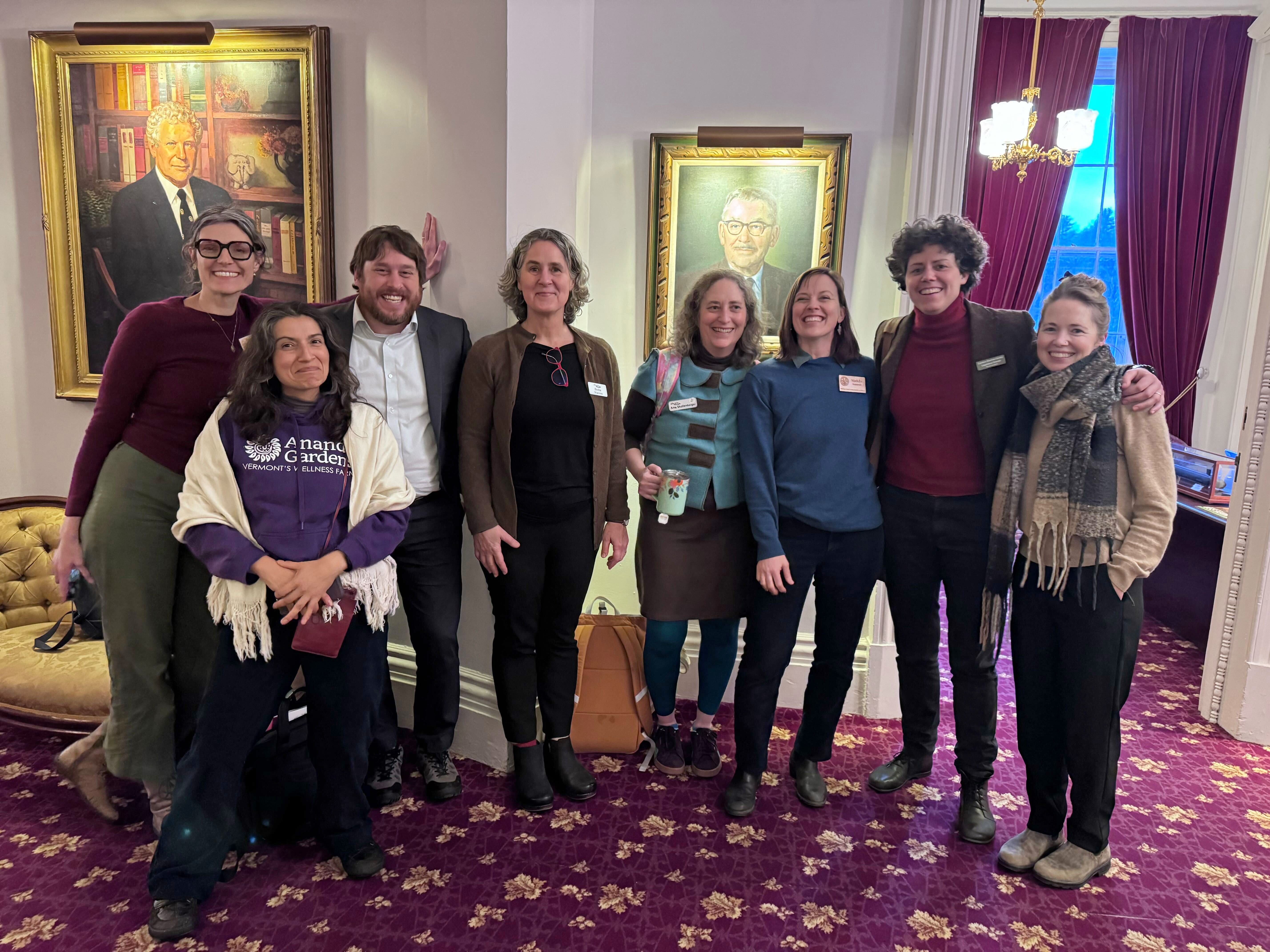NOFA-VT Grassroots Organizer Jess Hays Lucas, NOFA-VT farmer-member Melisa Oliva, Farm to Plate Director Jake Claro, Vermont Food Security Roadmap Coalition Manager Becka Warren, Action Circles Political Stragetist Amy Shollenberger, NOFA-VT Policy & Organizing Director Maddie Kempner, Rural Vermont Legislative Director Caroline Gordon, and Vermont Farm Fund Manager Kristin Blodgett stand arm in arm smiling at the Vermont State House after a successful day of lobbying in support of S.60