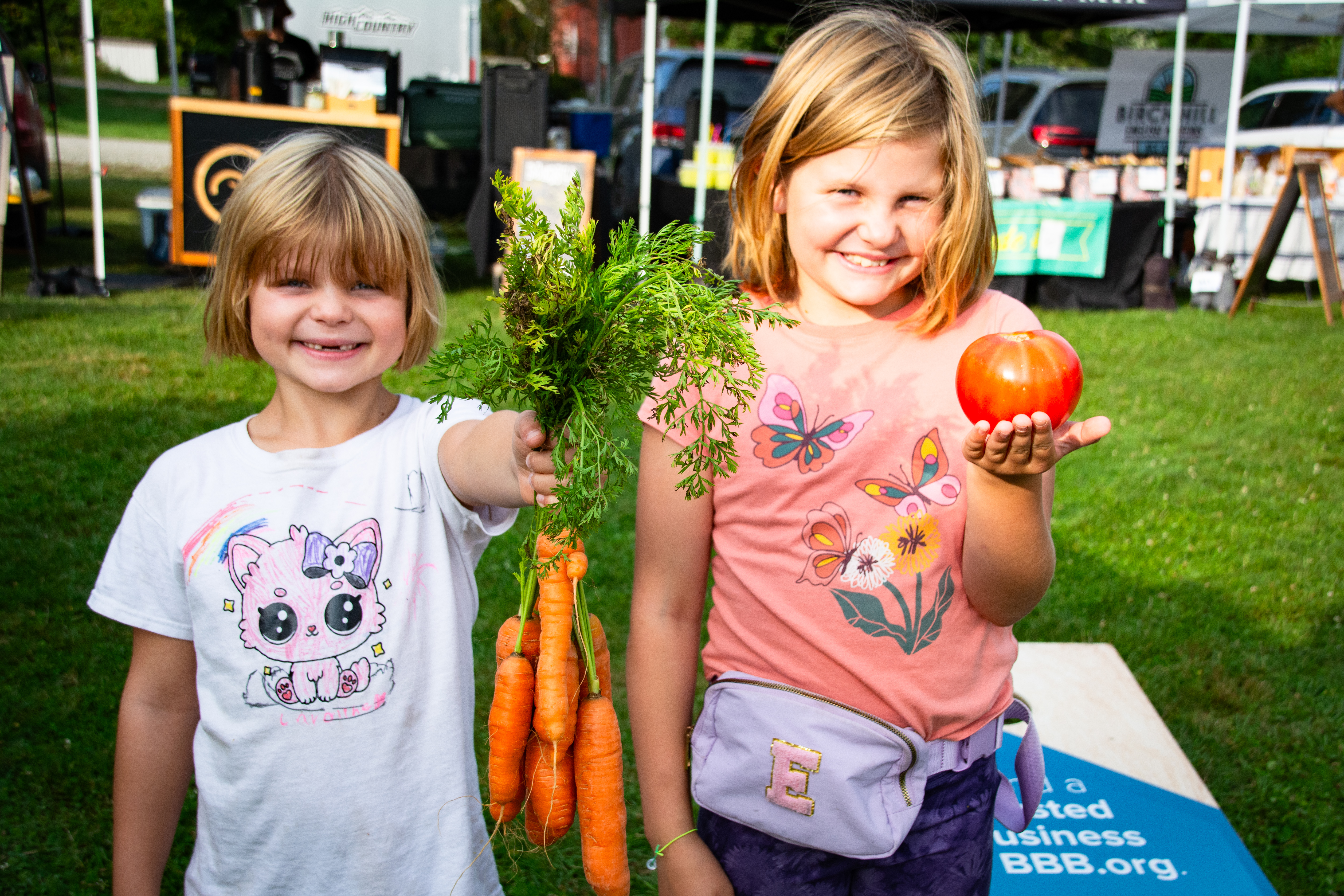 Two small children smile while holding up carrots (left) and a tomato (right) at a Vermont farmers market