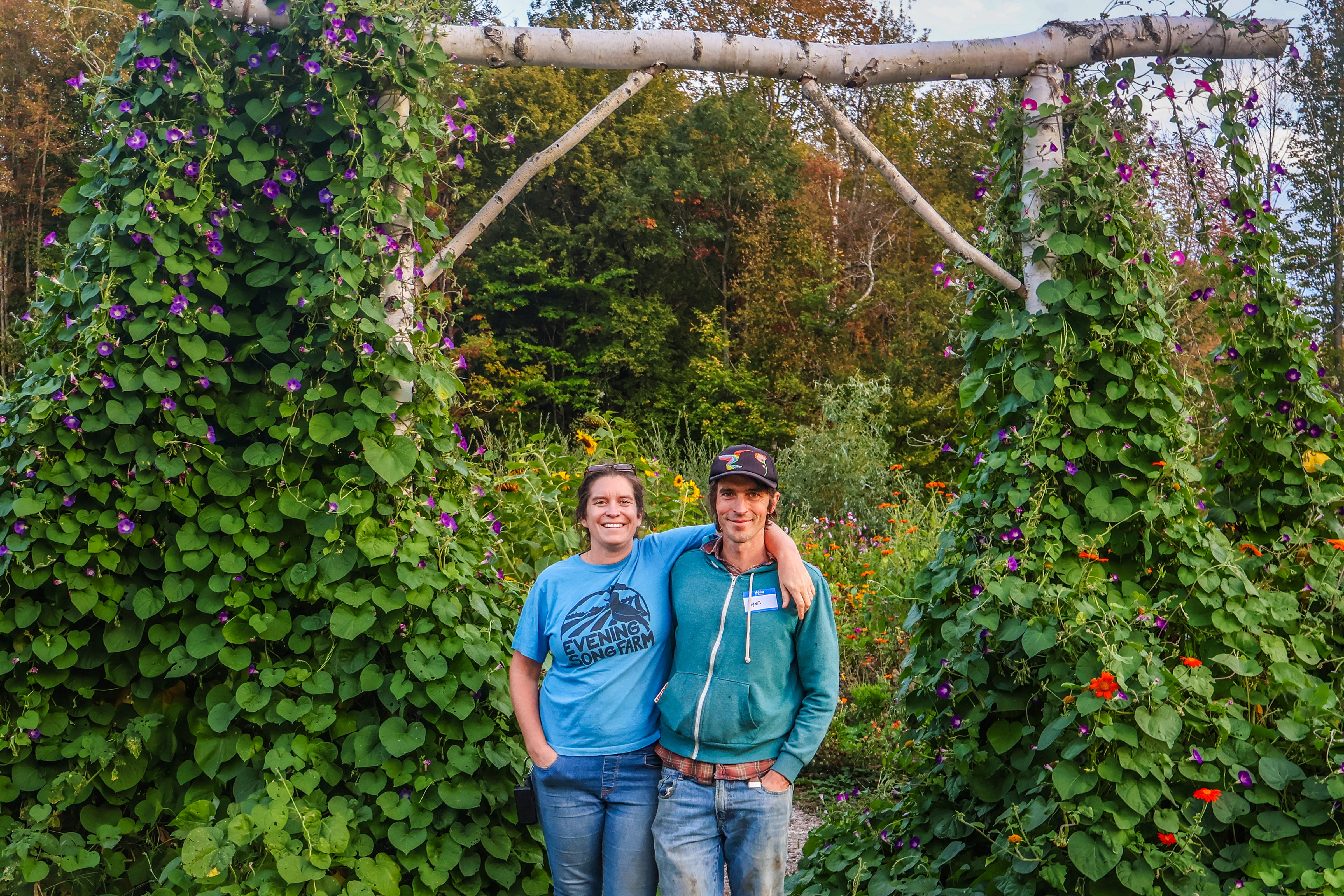Kara and Ryan Fitzbeauchamp of Evening Song Farm are pictured arm in arm, smiling, under a birch arbor covered in flowering morning glory vines, with their lush farm fields in the background