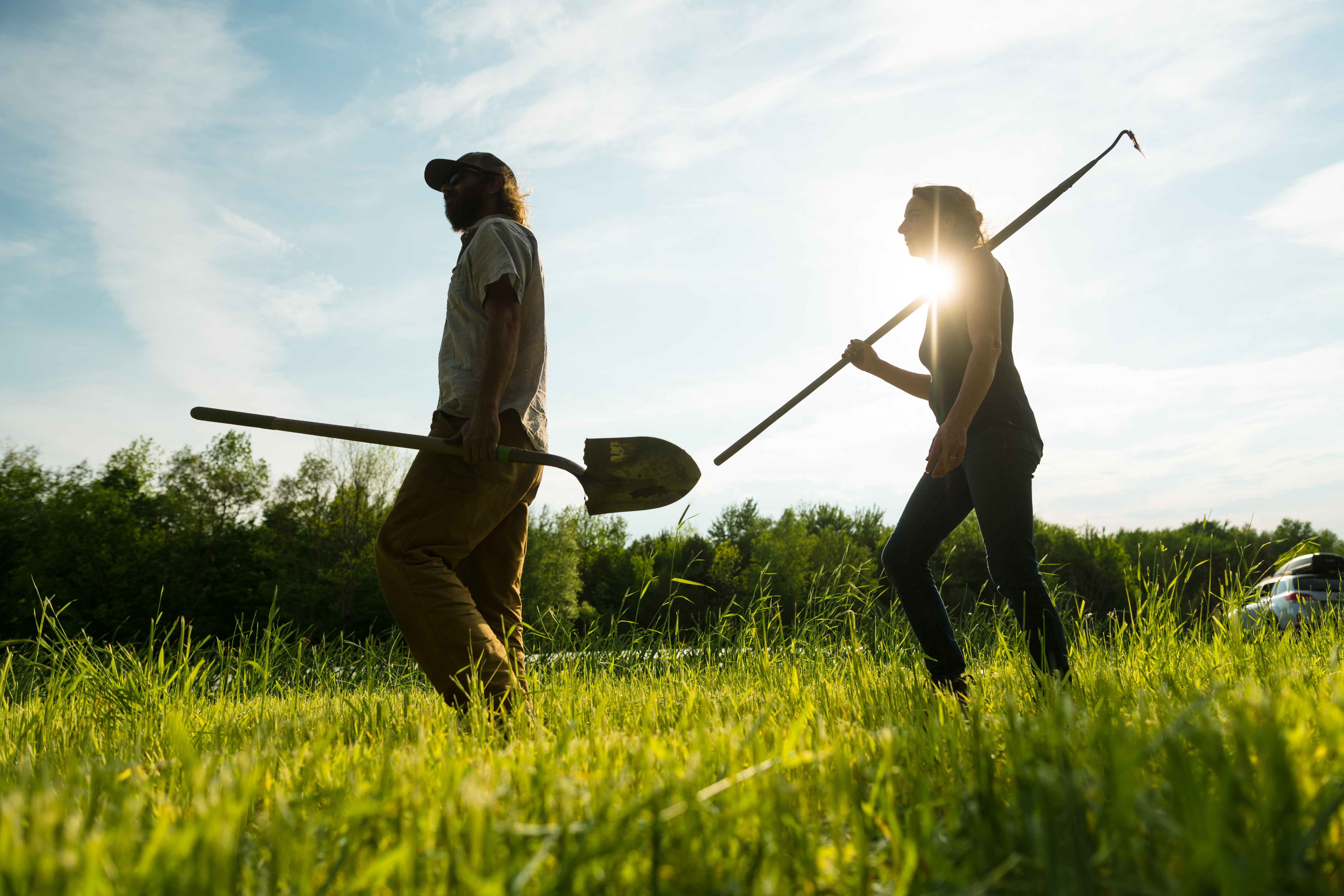Two farmers from Knee Deep Farm walking through their farm field holding a hoe and a shovel with a bright sun behind them, causing them to appear as silhouettes.