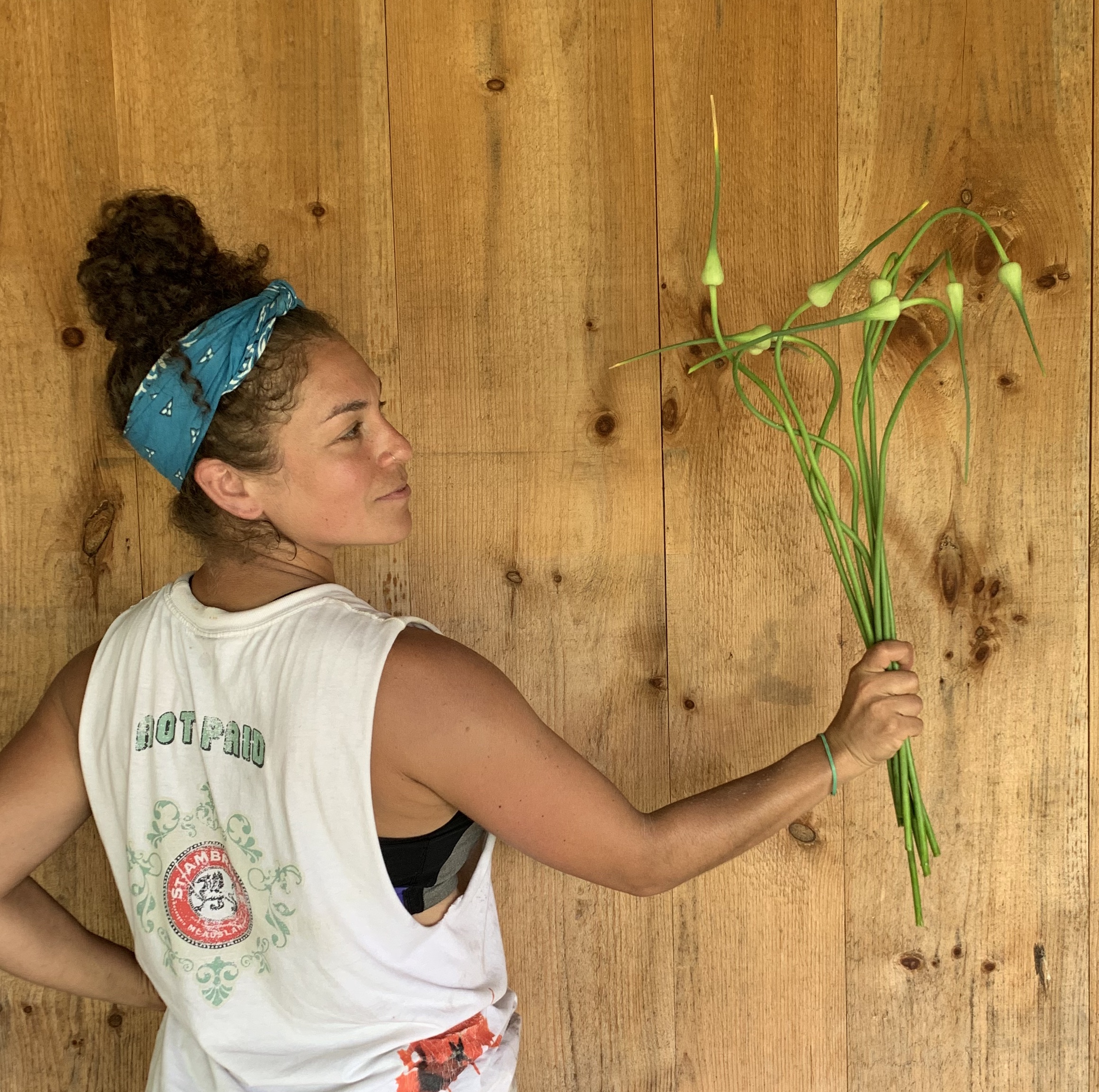 Amara Watkin-Anson from La Finca Cooperative stands with one hand on her hip, and the other hand holding a bundle of fresh garlic scapes