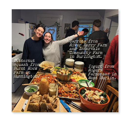A group of people mill around a kitchen with a spread of food on a table in the middle. Two people face the camera, smiling with their arms out wide.