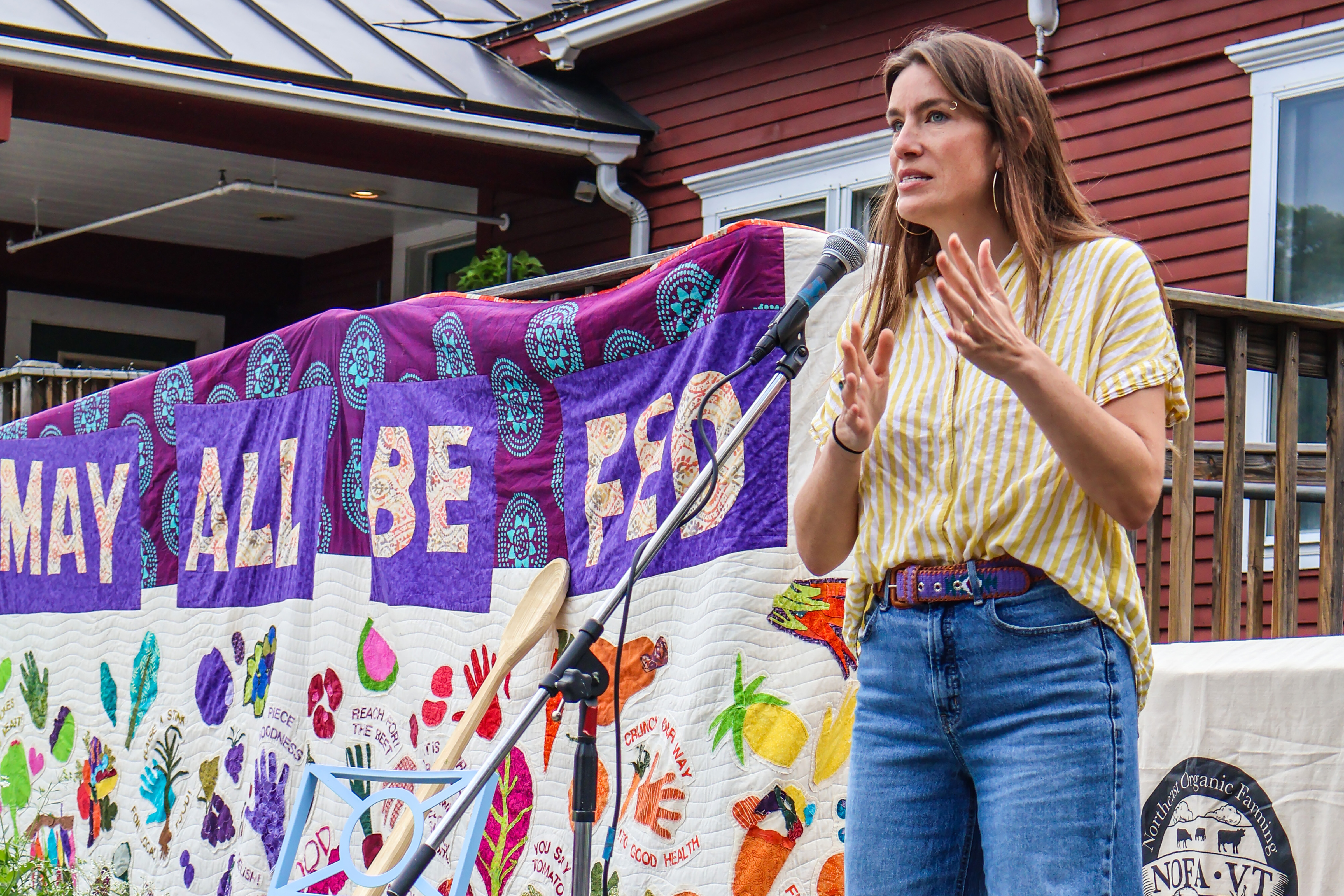 NOFA-VT Executive Director Grace Oedel addresses a crowd at the Upper Valley Food Co-op parking lot, with a banner behind her reading "May All Be Fed." Participants were gathered for an Upper Valley Farmer Day of Action in 2025.
