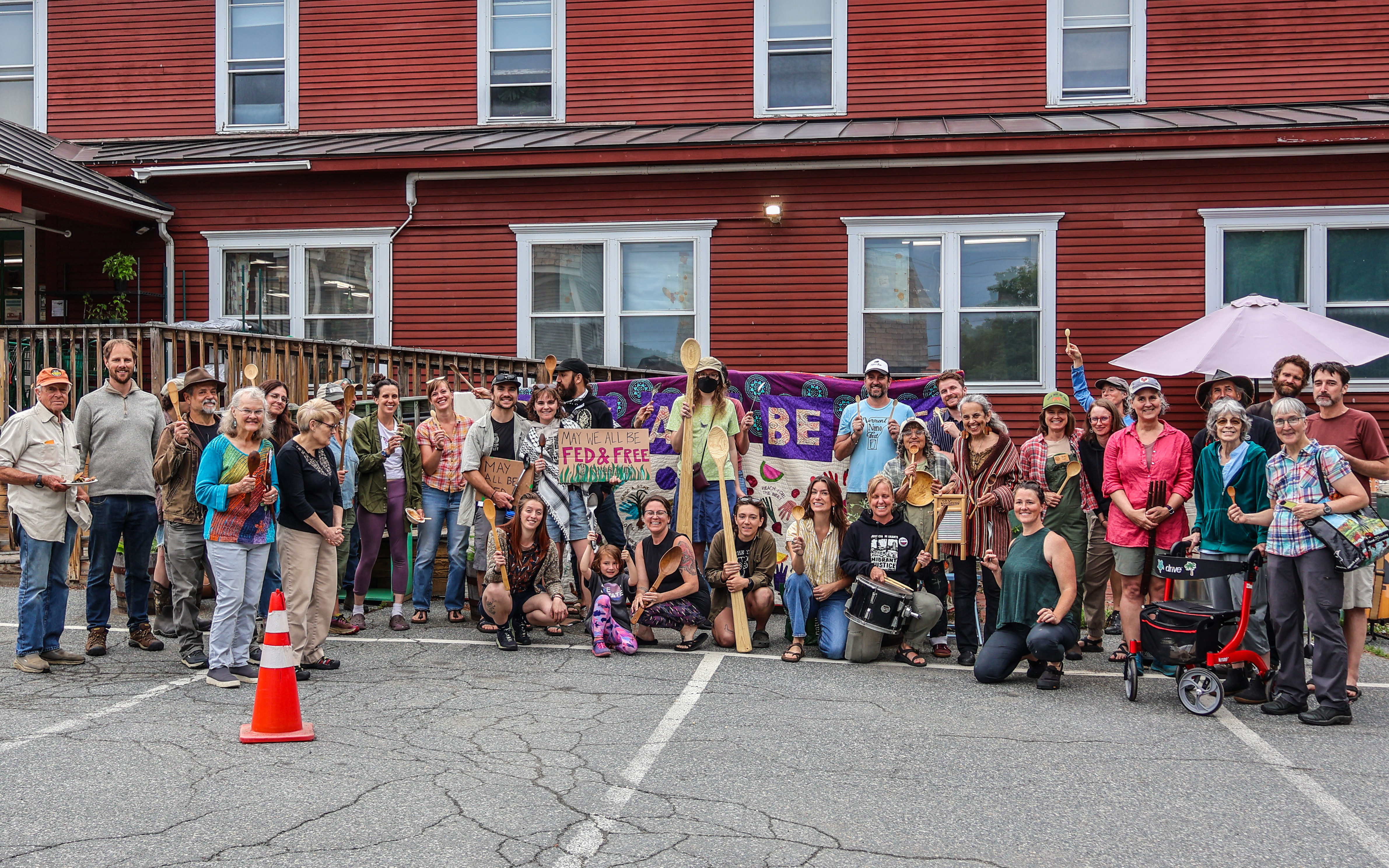 A large group of farmers, farmworkers, and food system advocates standing together in a parking lot at the Upper Valley Food Co-op, smiling, and holding signs in support of farmer and farmworker rights