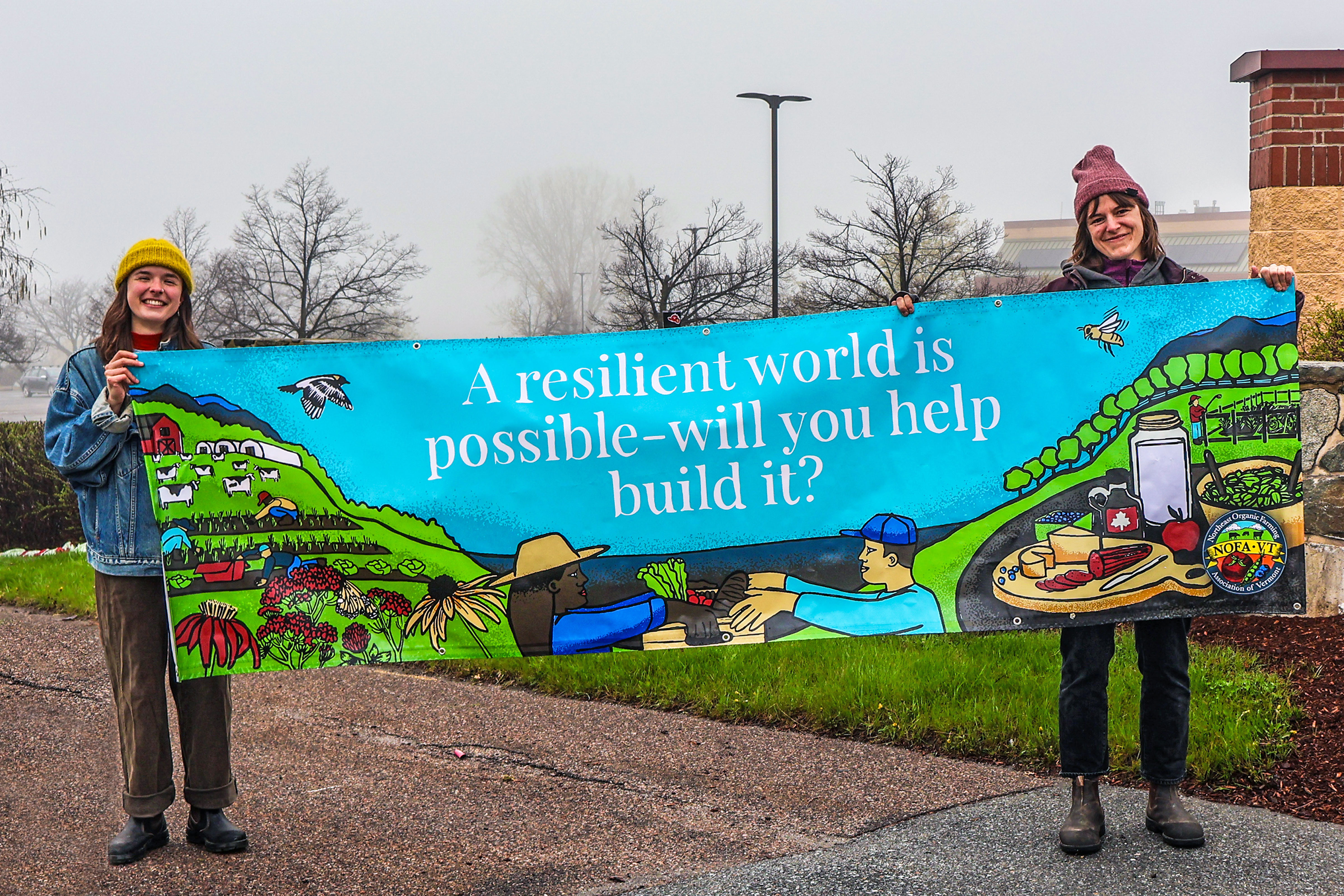 Two smiling NOFA-VT staff members holding a banner that reads "A resilient world is possible—will you help build it?"