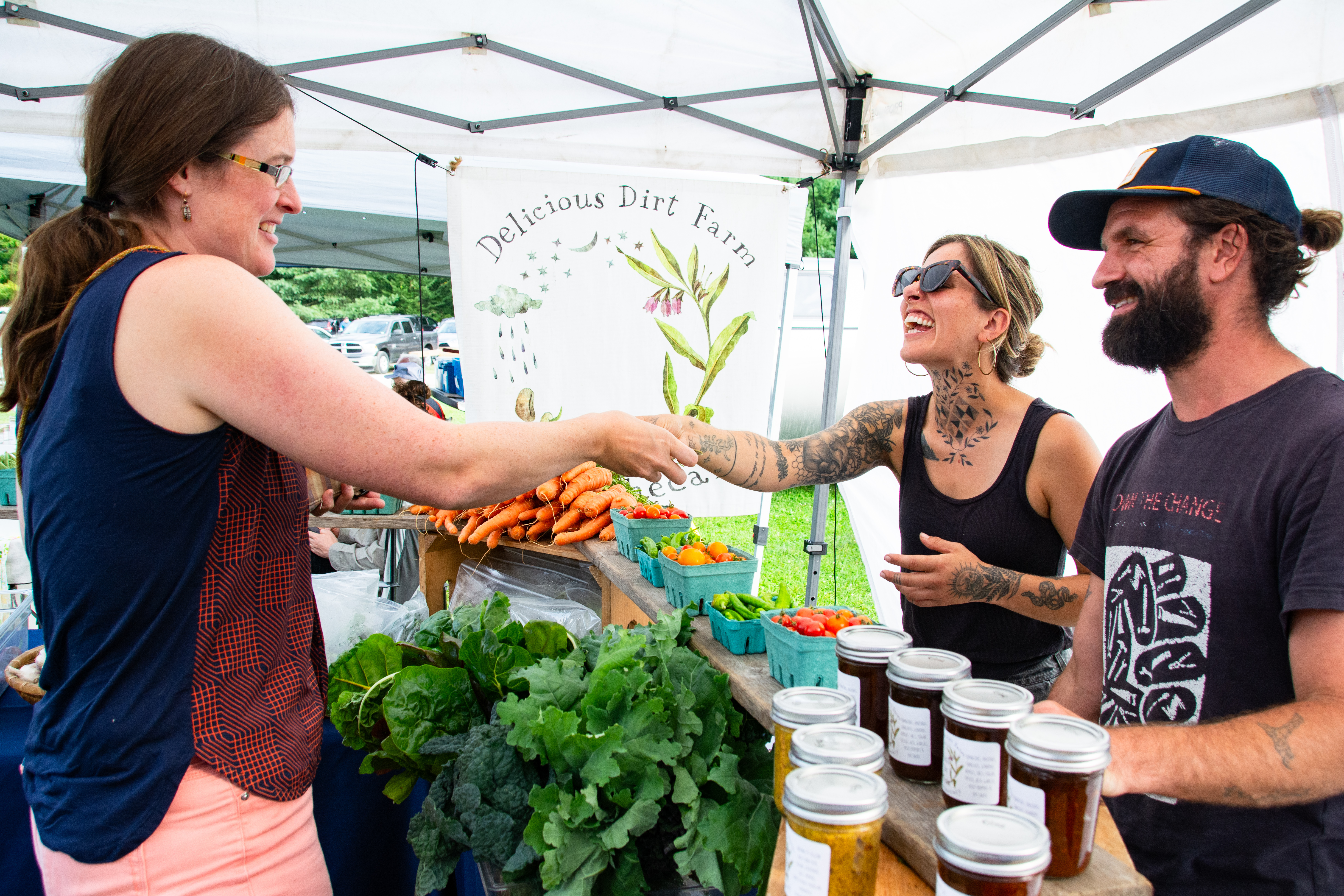 two smiling customers at a farmers market pass crop cash to a farmer from Delicious Dirt Farm