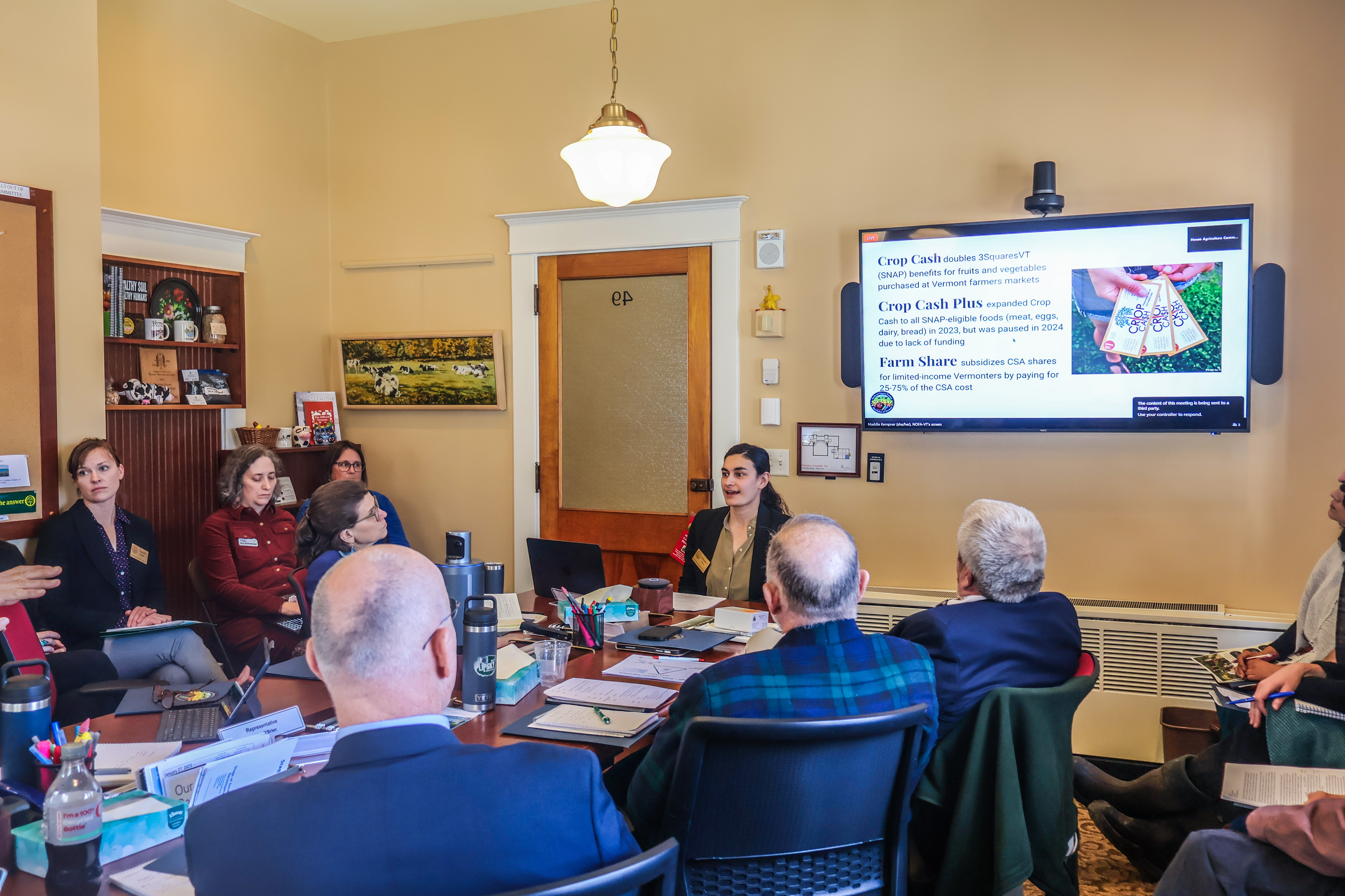 Johanna Doren, NOFA-VT Direct Markets and Local Food Access Director, provides testimony in support of NOFA-VT's state funding request to support Crop Cash, Crop Cash Plus, and Farm Share, as member of the Ag committee sit around a large table listing to Johanna's remarks.