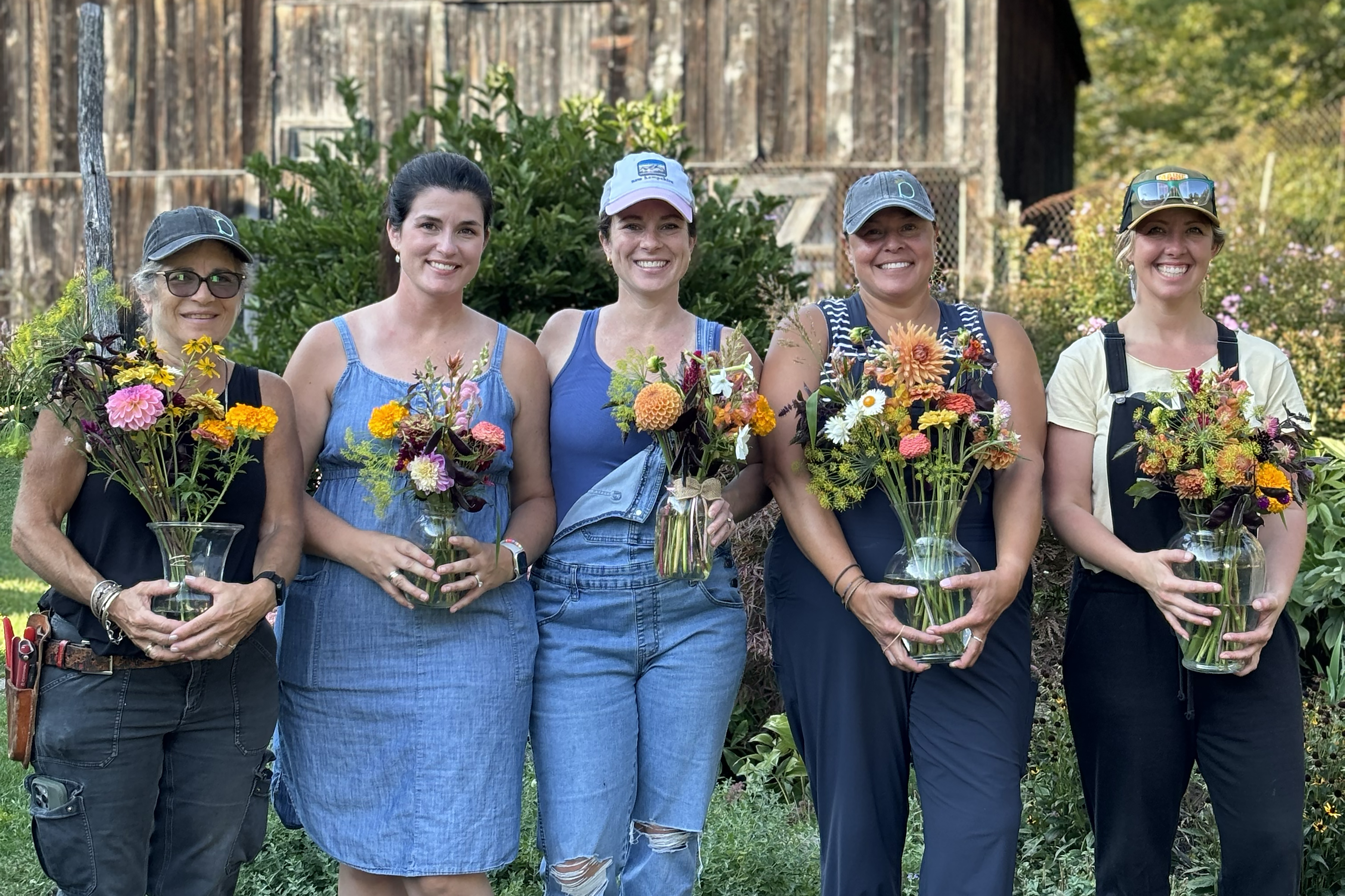 participants at a flower workshop at By Hand Farm stand smiling with their bouquets with a barn in the background