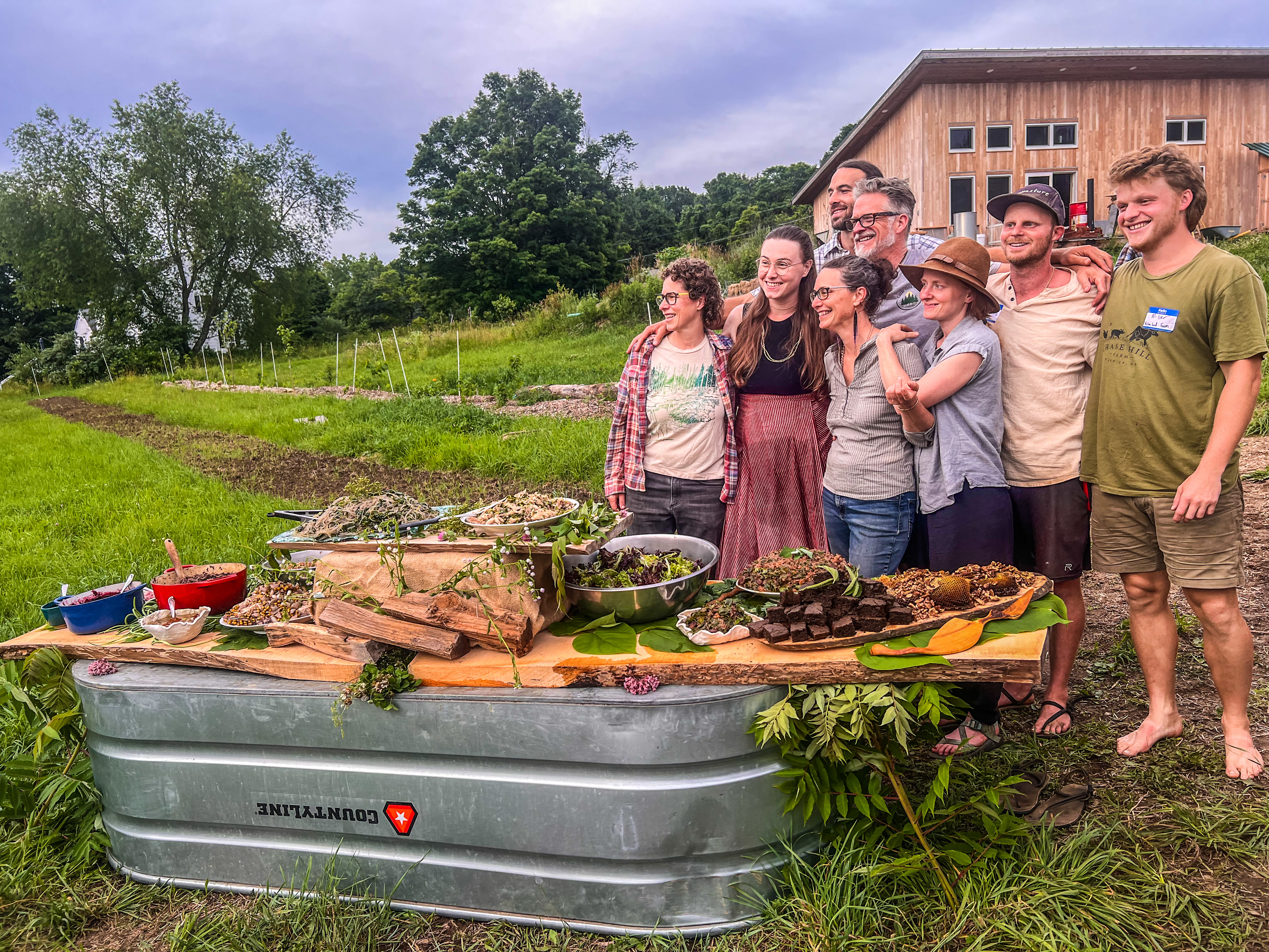 Participants from the regional agroforestry gathering stand smiling together next to a beautiful spread of perennial foods laid out on a plank atop a large galvanized wash tub. Crops are planted in the background.