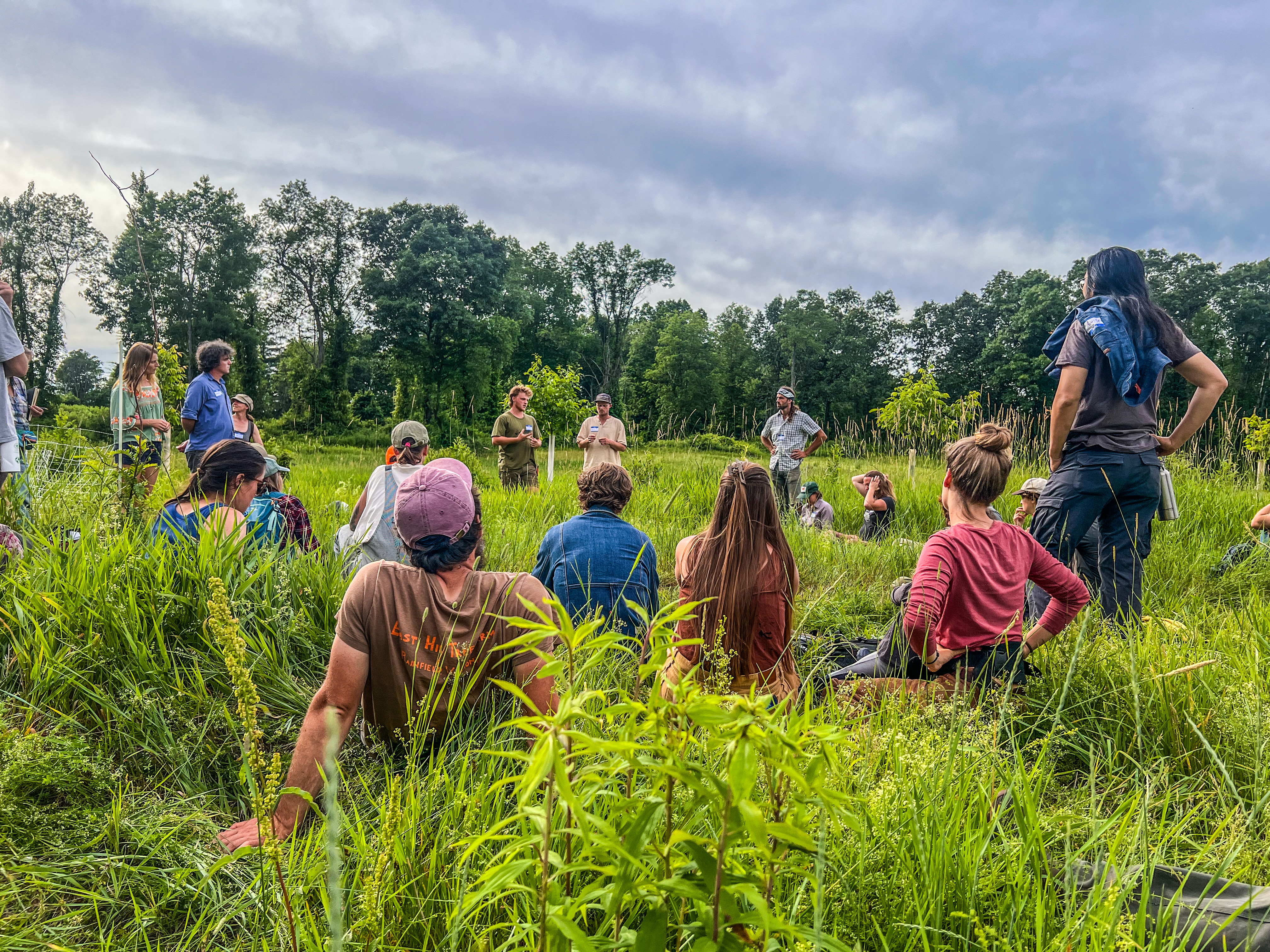 Participants from the regional agroforestry gathering are shown seated in a circle in a field discussing perennial agriculture