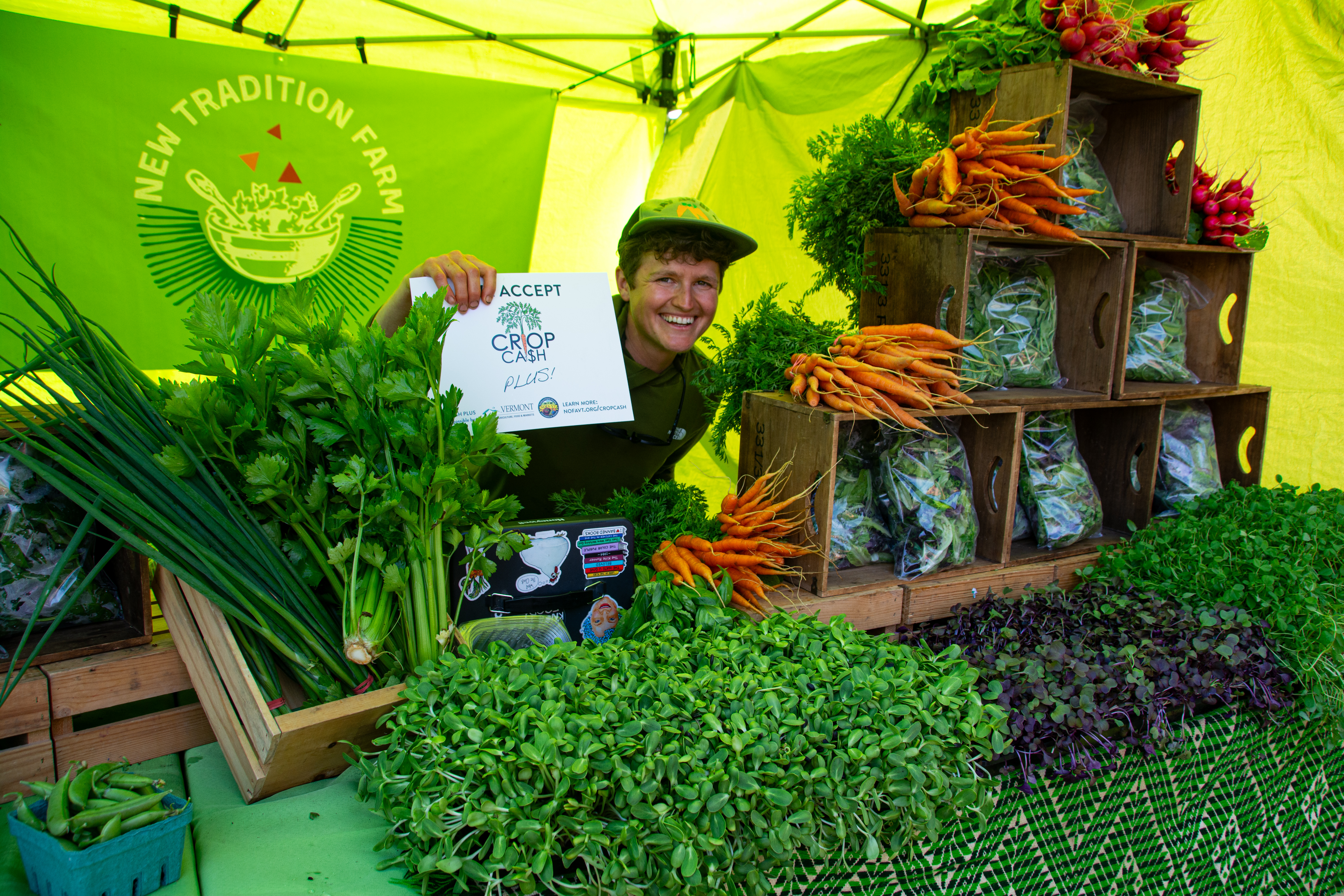 NOFA-VT Farmer Member Stoni Tomson of New Tradition Farm stands smiling behind their market booth, holding a sign reading "We Accept Crop Cash and Crop Cash Plus!"