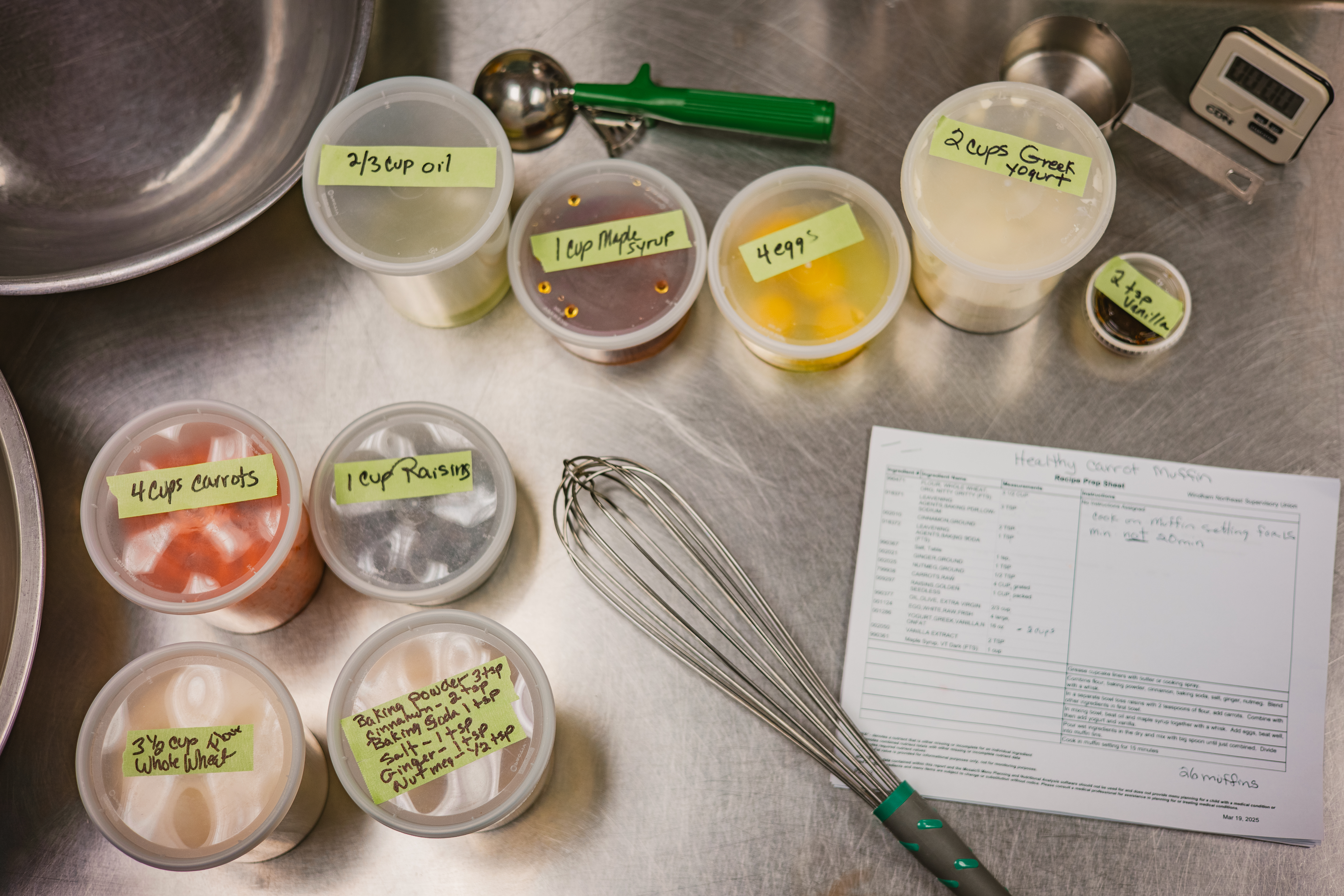 A collection of ingredients and cooking utensils spread out on a stainless steel prep table in a school cafeteria
