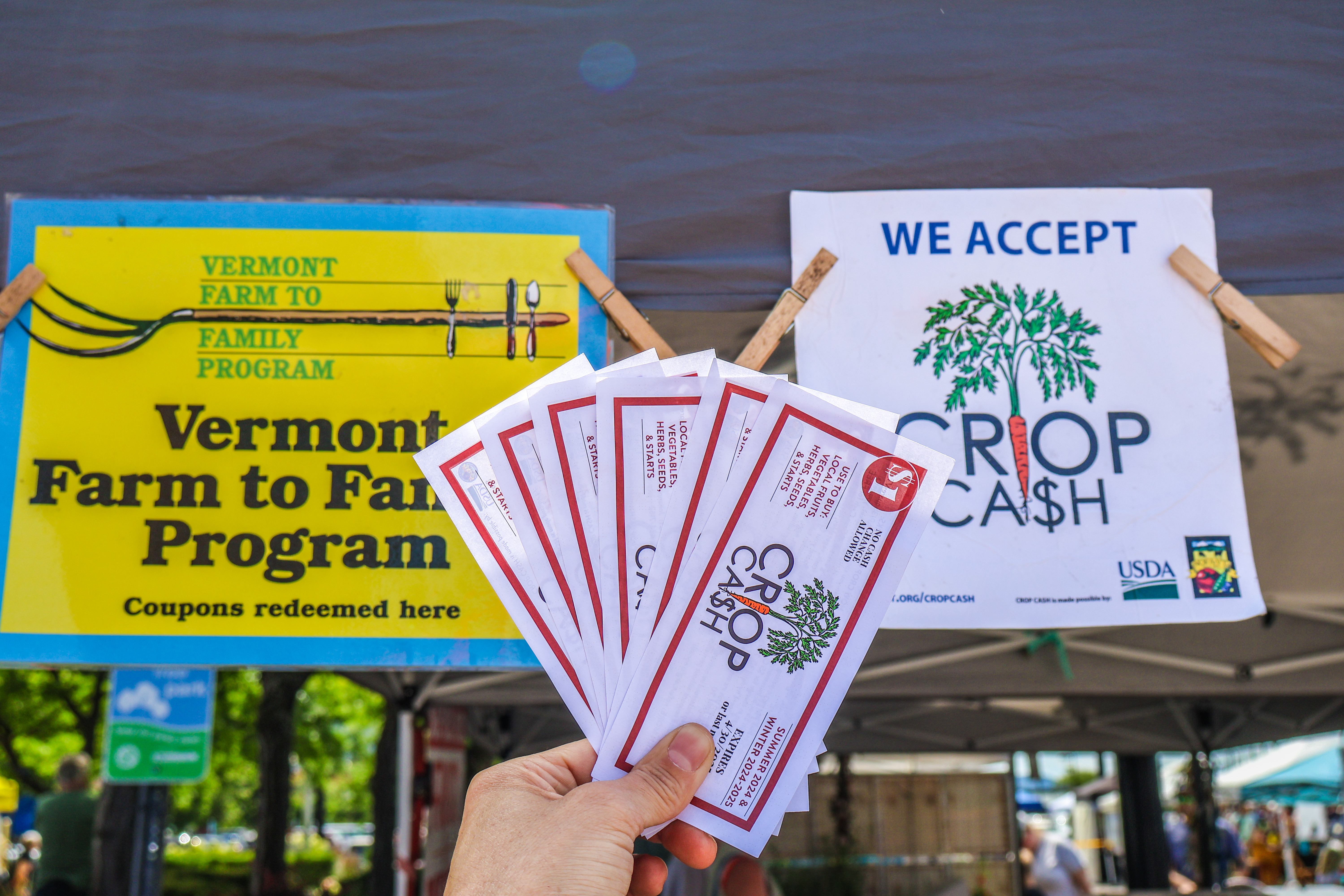 Crop Cash coupons fanned out in front to two farmers markets signs reading "Vermont Farm to Family Program Coupons redeemed here" and "We Accept Crop Cash"