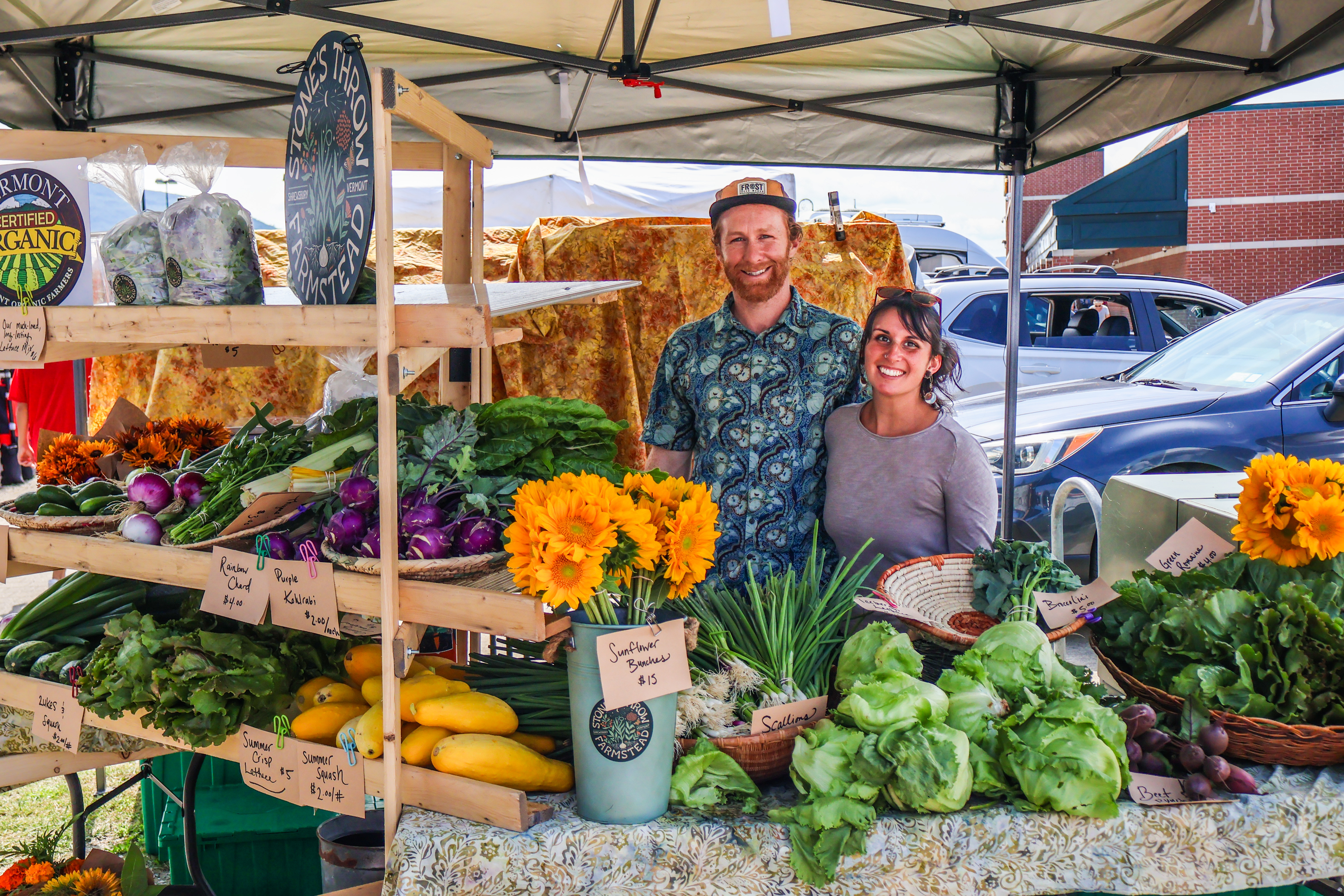 Connor and Jess stand smiling behind an abundant spread of colorful flowers and veggies at their table at the Vermont Farmers Market in Rutland
