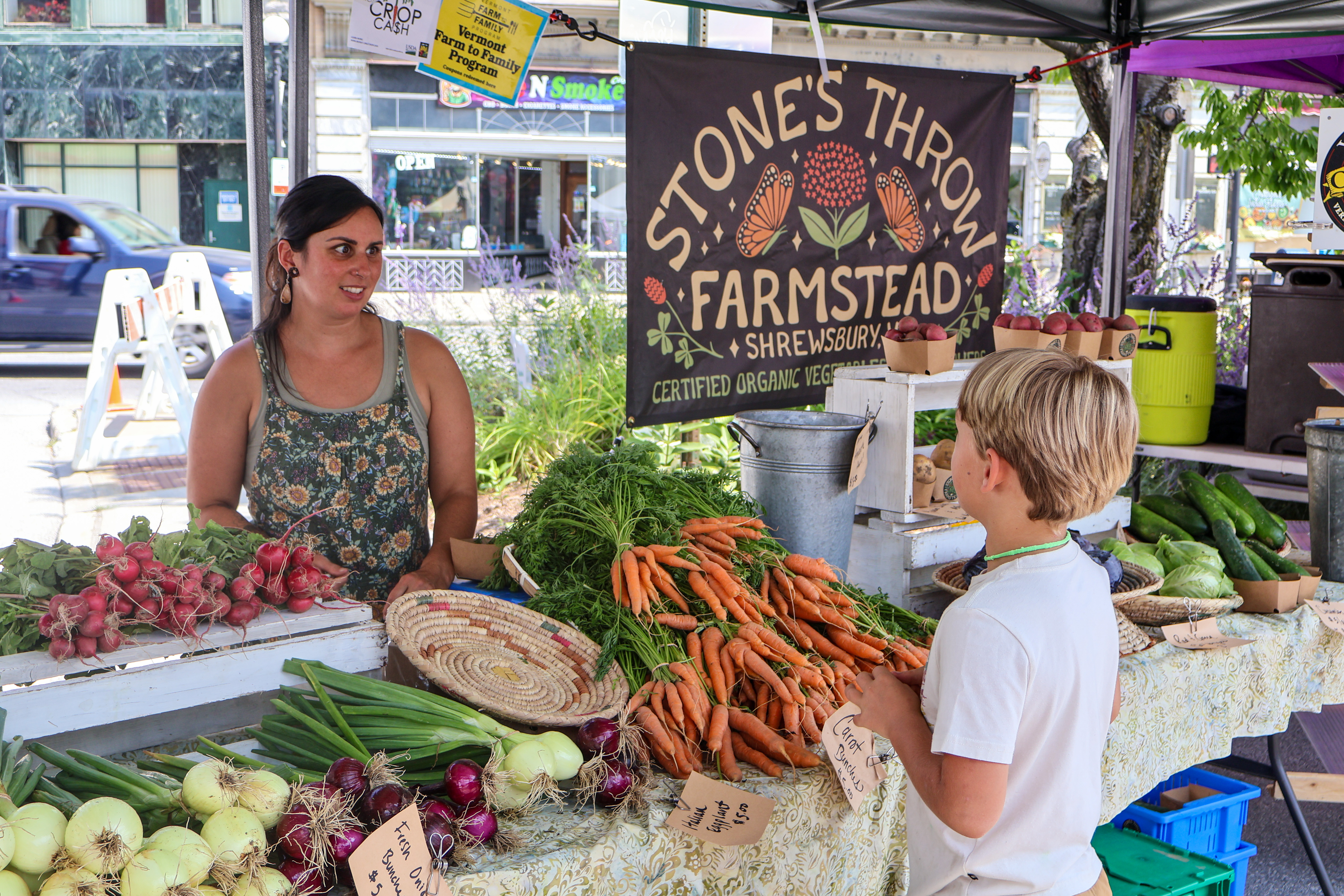 Farmer Jessica Purks of Stone's Throw Farmstead chats with a young customer over a table of colorful organic produce at the Vermont Farmers Market