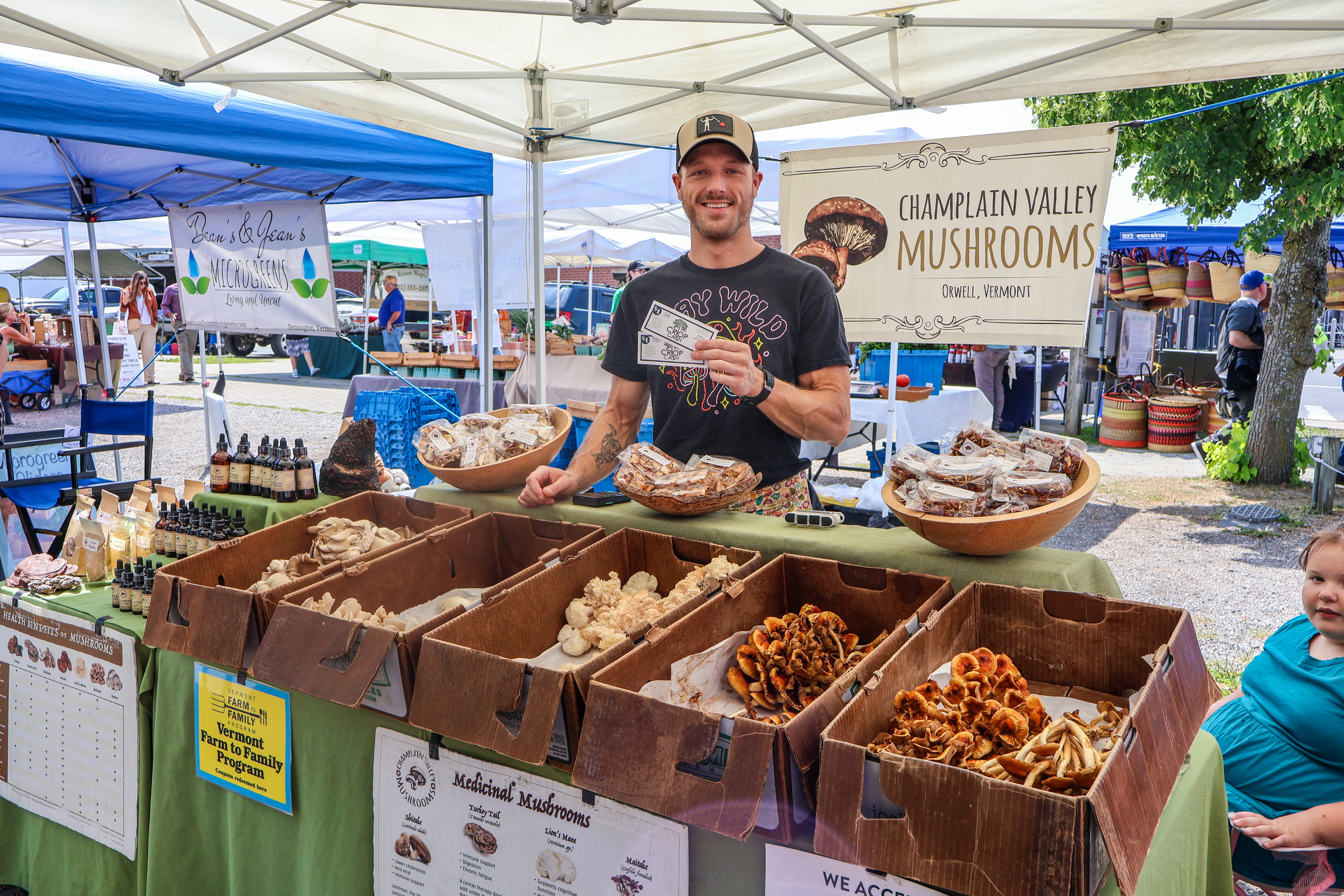 A representative from Champlain Valley Mushrooms stands smiling behind his farmers market table holding several crop cash coupons that customers used to purchase his products