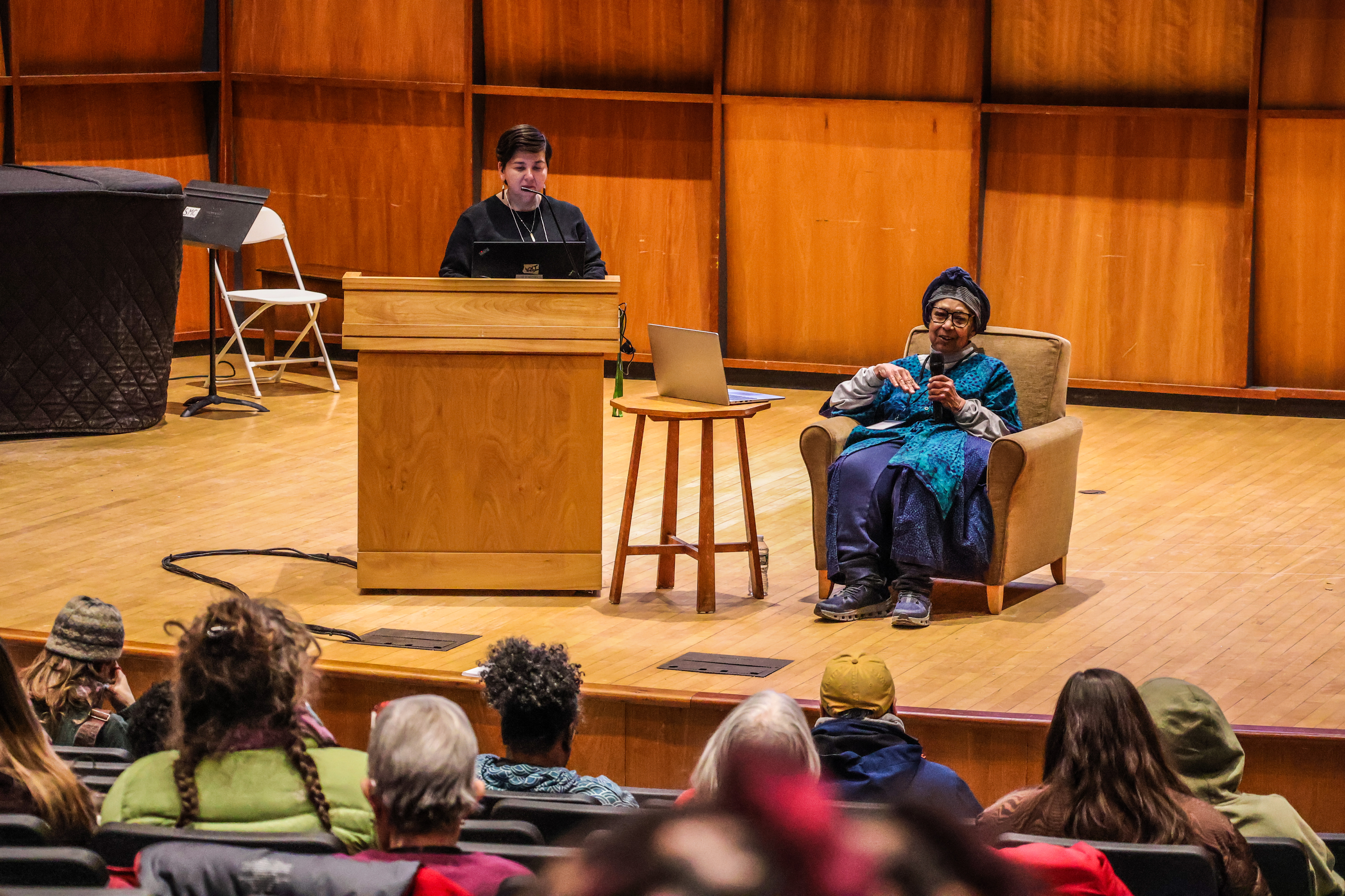 Alexis Yamashita and Ira Wallace are pictured on stage in a large auditorium at Saint Michael's College, leading a workshop as part of NOFA-VT's winter conference, with participants in several rows of filled seats enjoying the presentation