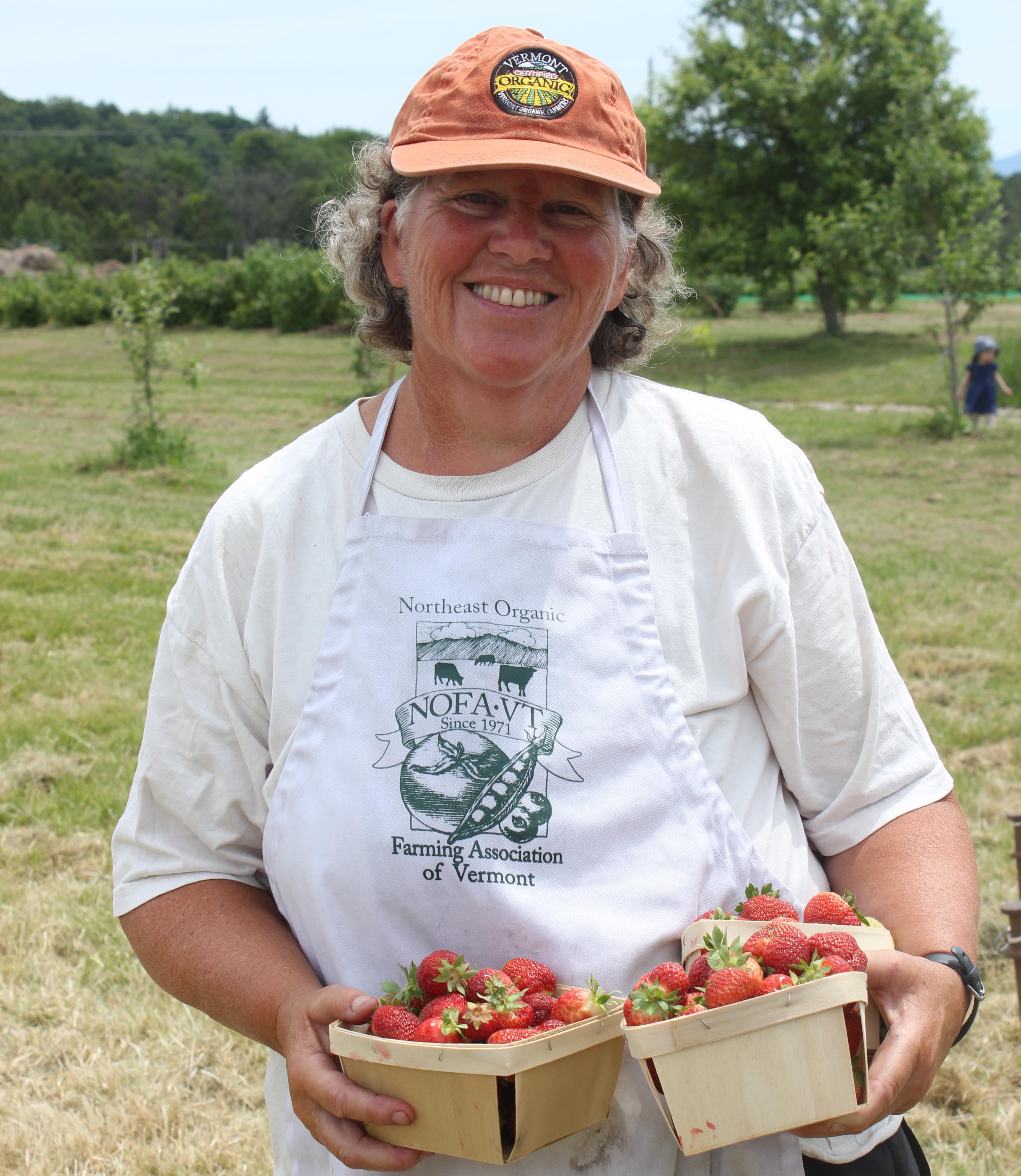 Enid stands wearing a VOF hat and a NOFA-VT apron, holding a carton of strawberries and smiling.