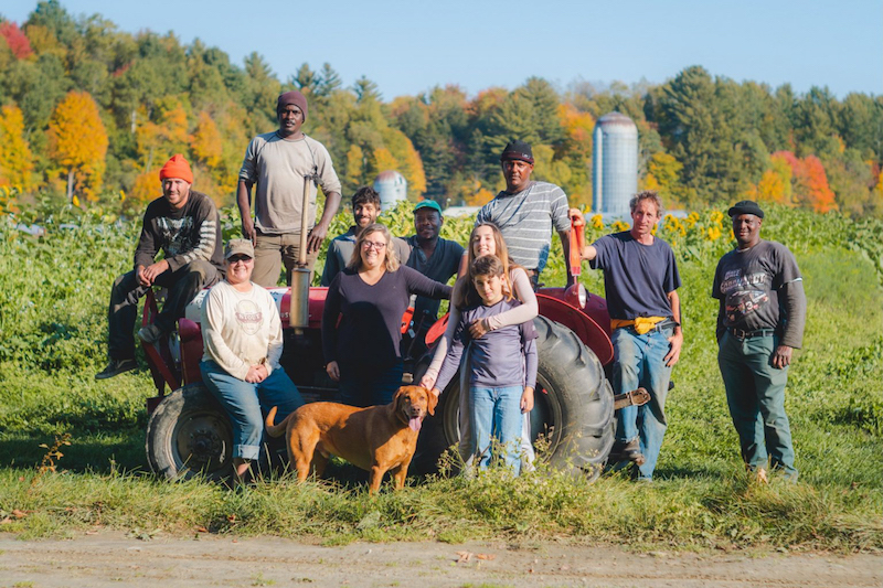 Roughly ten people stand smiling in front of a small farm field