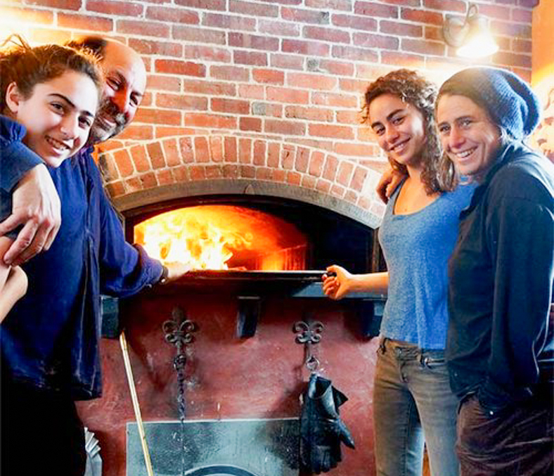 Four people stand, embracing in front of a wood fired oven