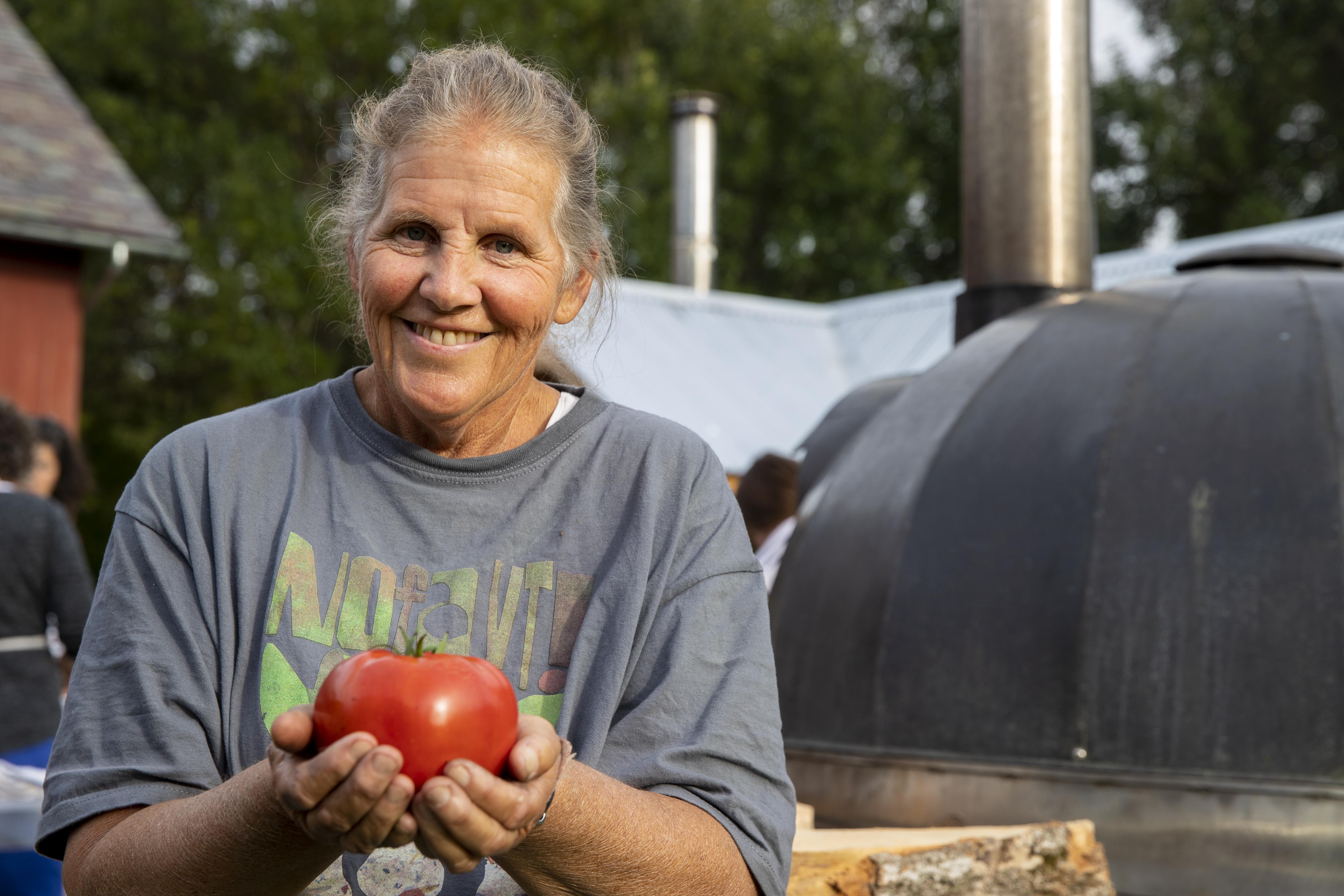Enid Wonnacott stands outside in a NOFA-VT tee-shirt holding a tomato out to the camera and smiling. A woodfire pizza oven is in the background.