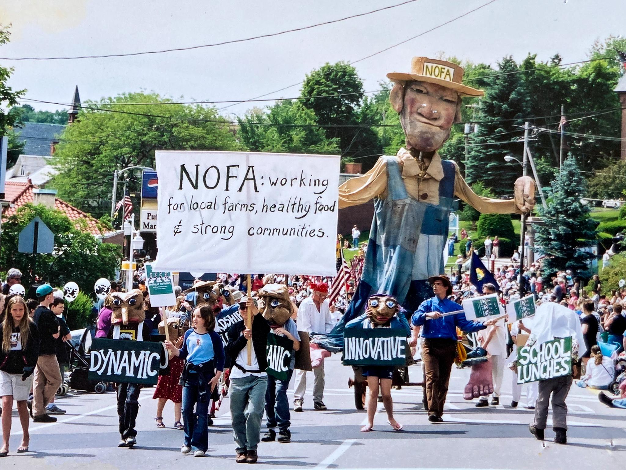 People walk down a street holding hand painted signs that say "dynamic", "innovative", "school lunches", and "NOFA: Working for local farms, healthy food, and strong communities". In the crown is also a 20 foot tall puppet wearing a hat that says "NOFA". 