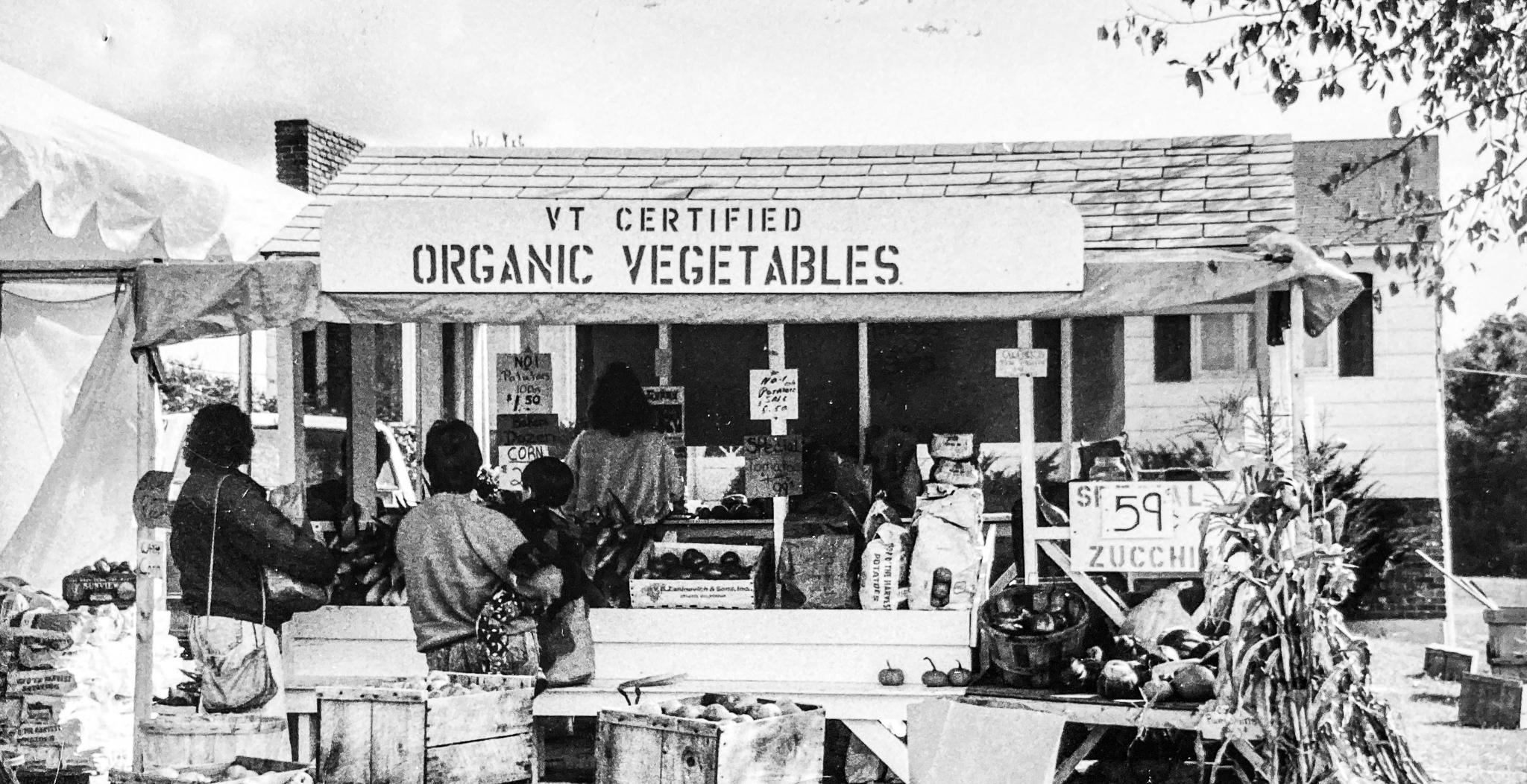 Black and white photo of people standing at a farm stand. The farm stand has a hand-stenciled sign above it that reads "VT Certified Organic Vegetables".