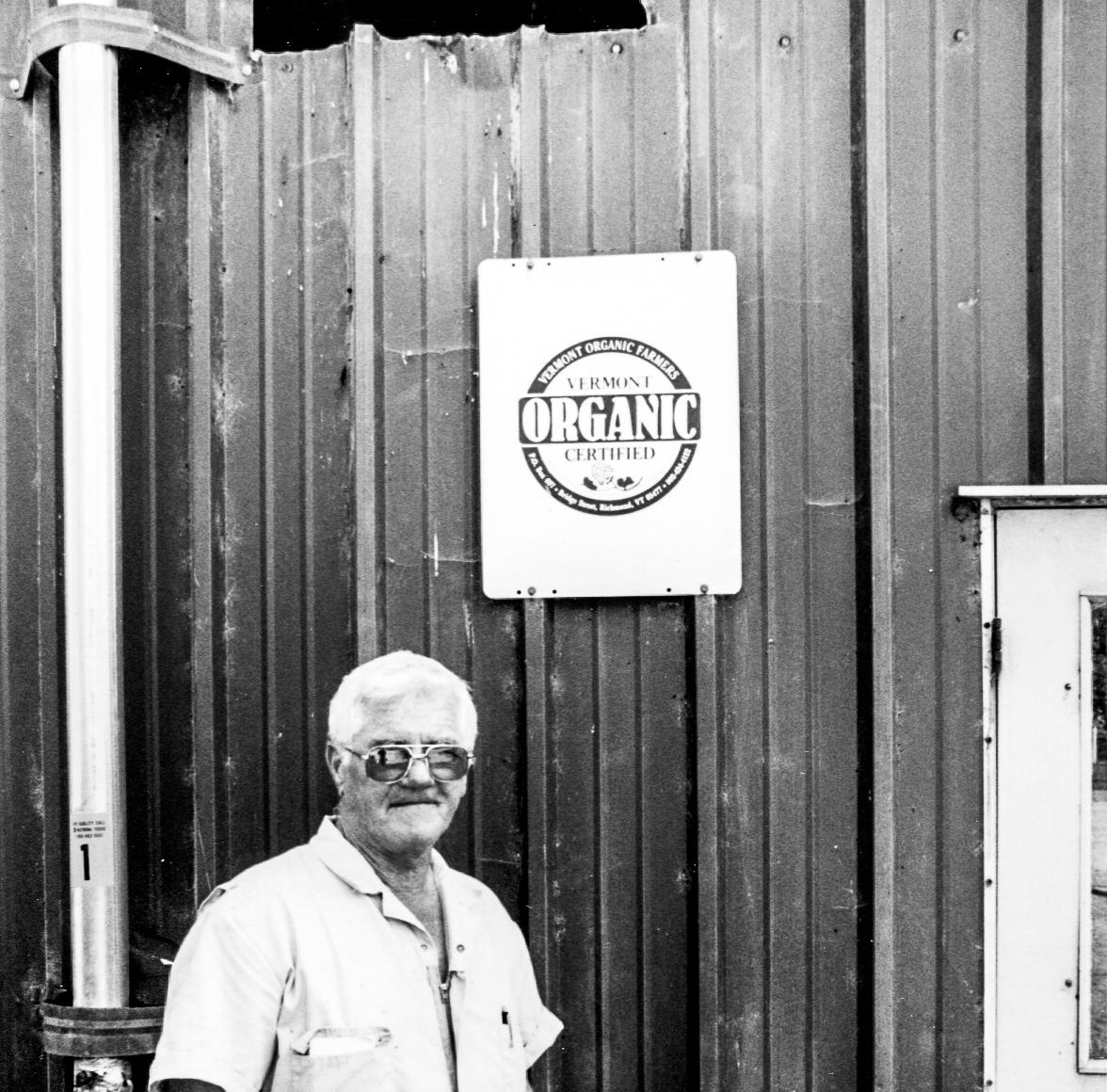 Black and white photo of a man standing in front of a barn that has a sign showing an old VOF logo.