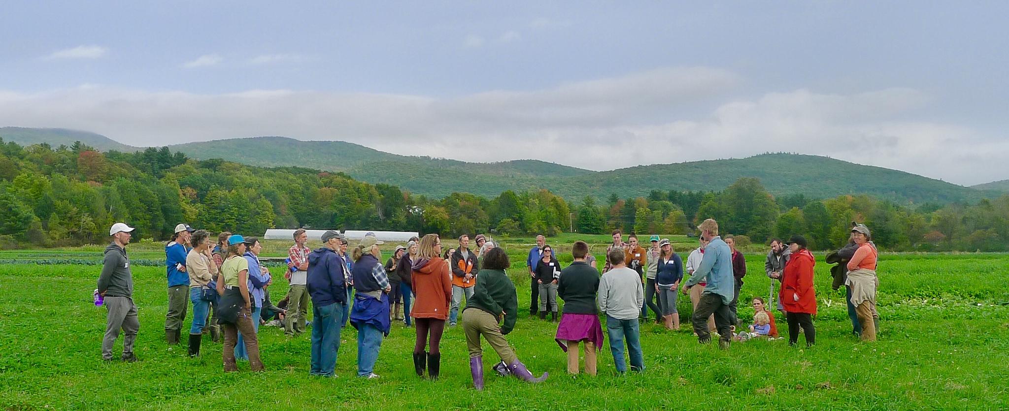 A large group of people stand in a circle in a grassy field with rolling hills in the background.