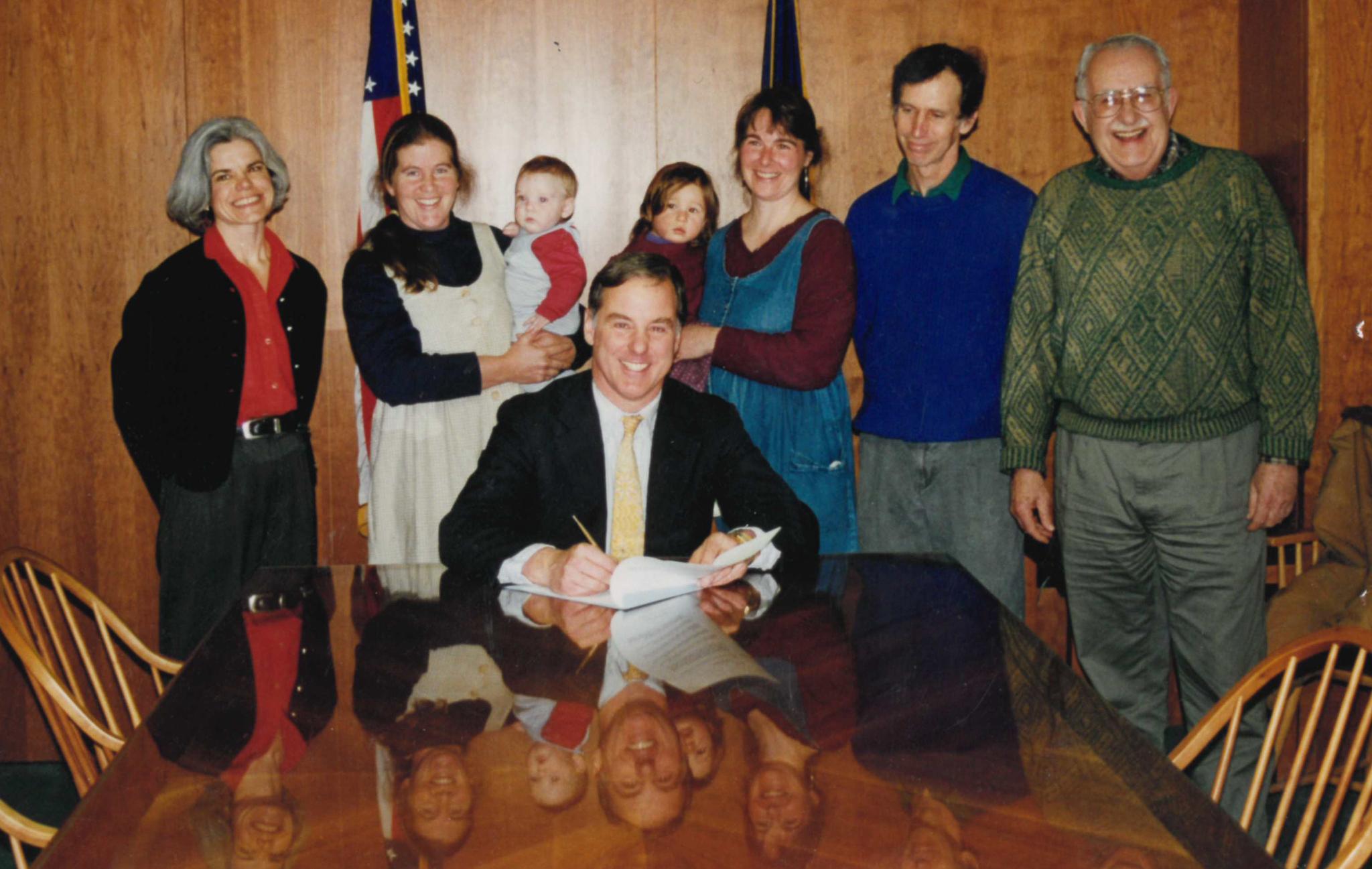 A group of five people (including former NOFA-VT Executive Director Enid Wonnacott and current NOFA-VT Director of Finance Kirsten Bower, both holding babies on their hips) stand behind former Vermont governor Howard Dean. Everyone is smiling at the camera, including Governor Dean, who is also poised to sign some papers.  