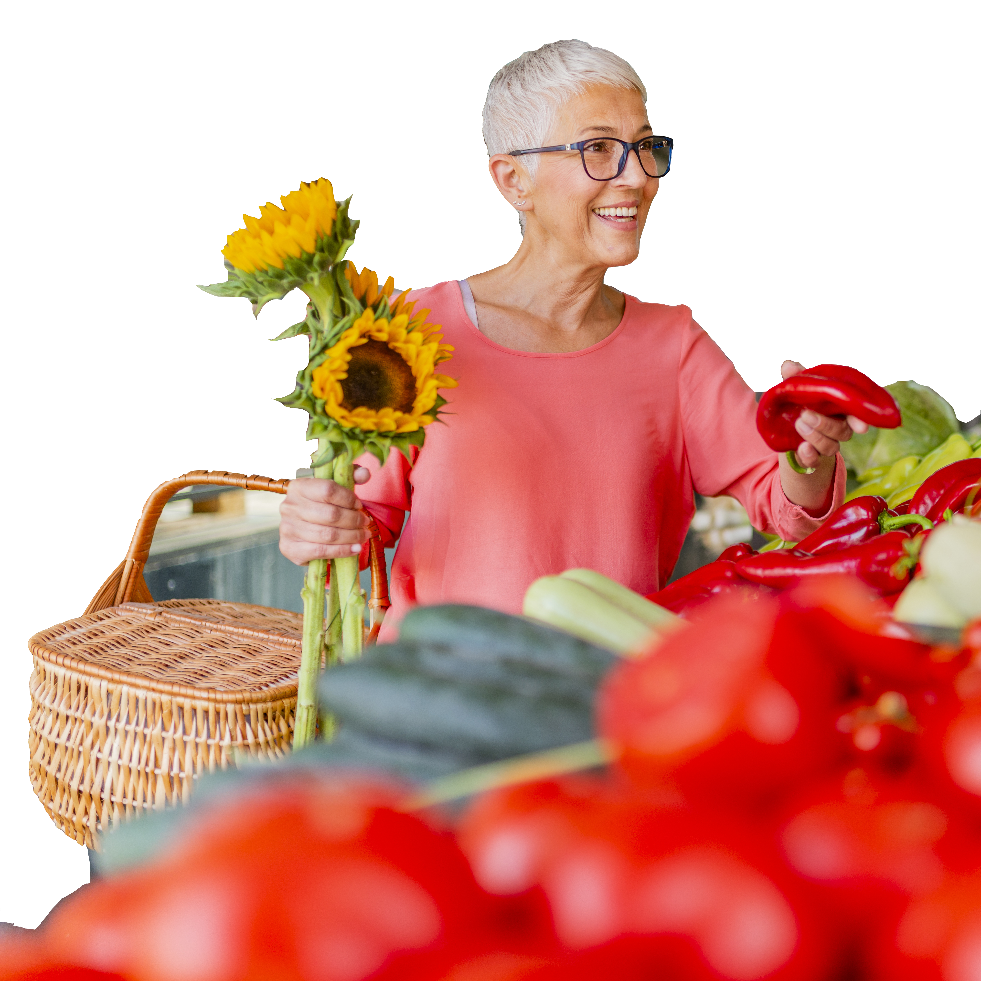 An older woman is smiling and holding a basket, sunflowers, and a red pepper.