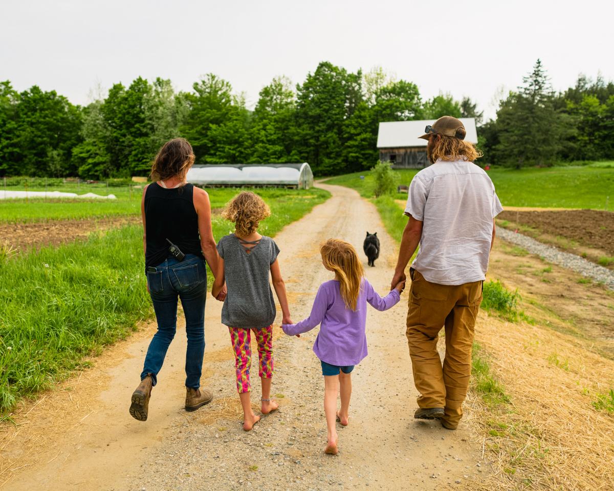 Two adults hold hands with their two children as they walk down a farm road
