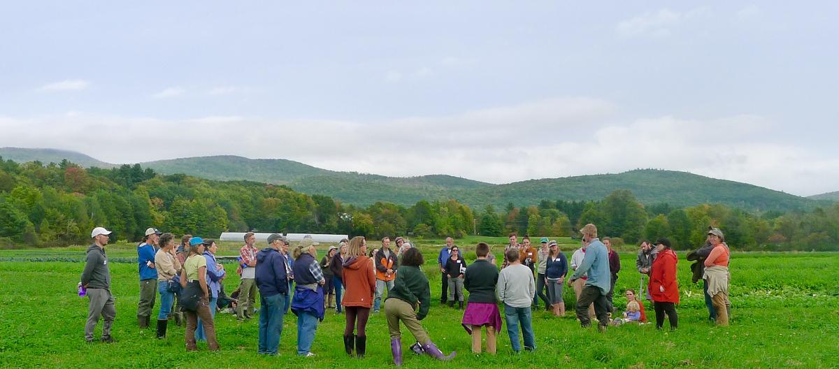 A group of roughly twenty people stand in a large circle in a grassy field, facing each other.